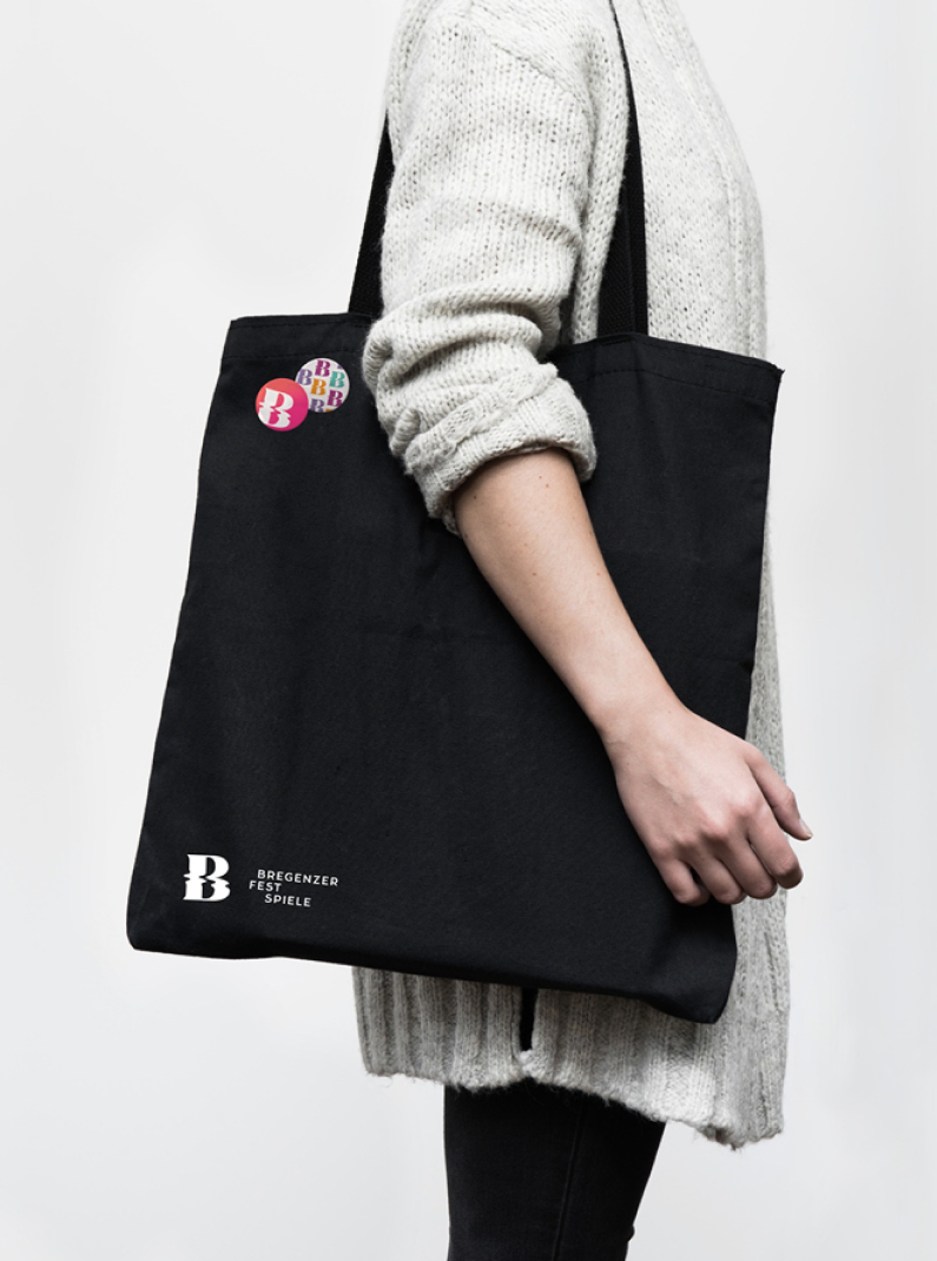 Eine Person hält eine schwarze Stofftasche der Bregenzer Festspiele mit Buttons vor einem weißen Hintergrund.