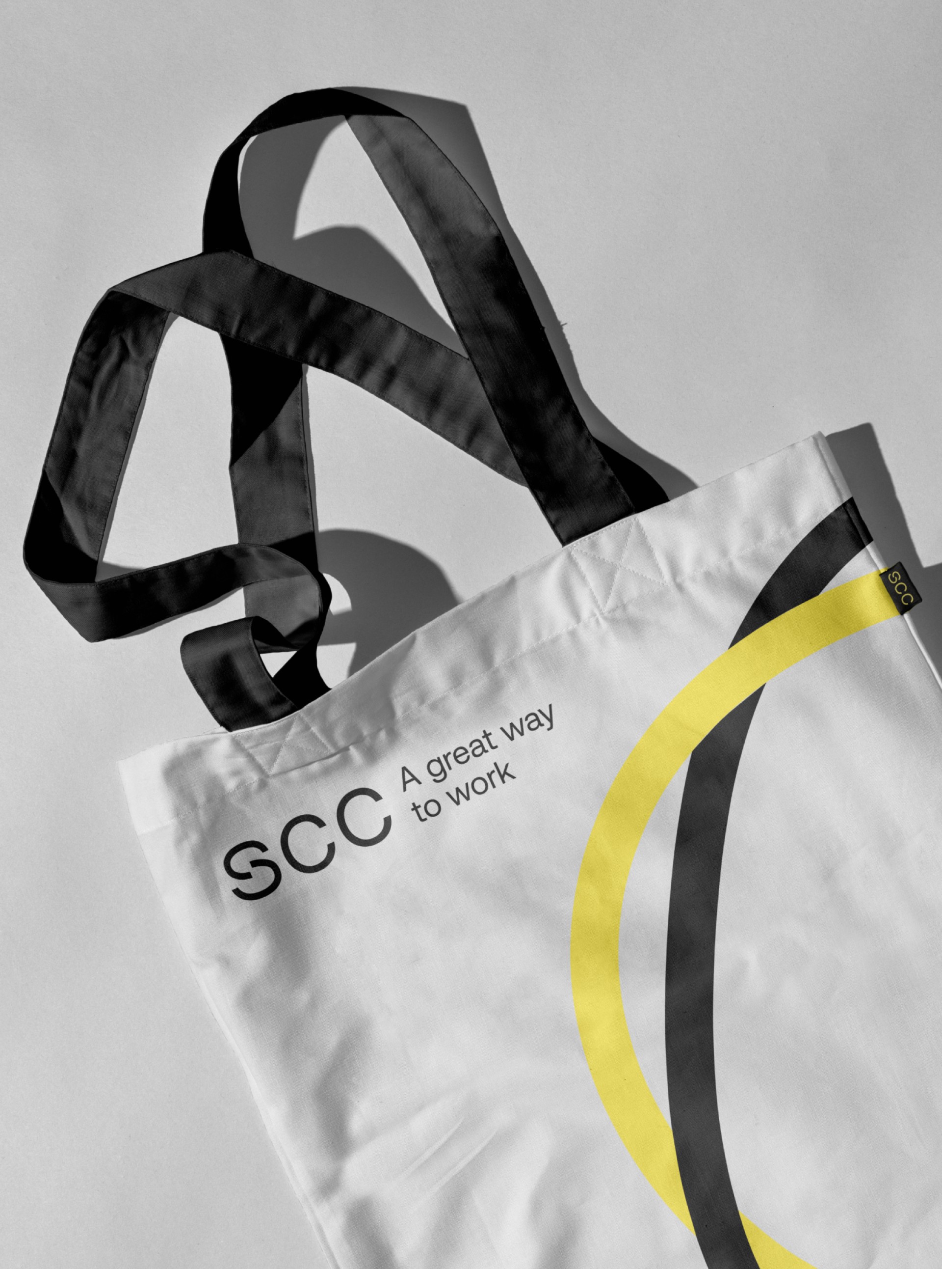 Eine weiße Einkaufstasche mit schwarzen Trägern, dem Text "SCC A great way to work" und einem gelb-schwarzen Kreisdesign.