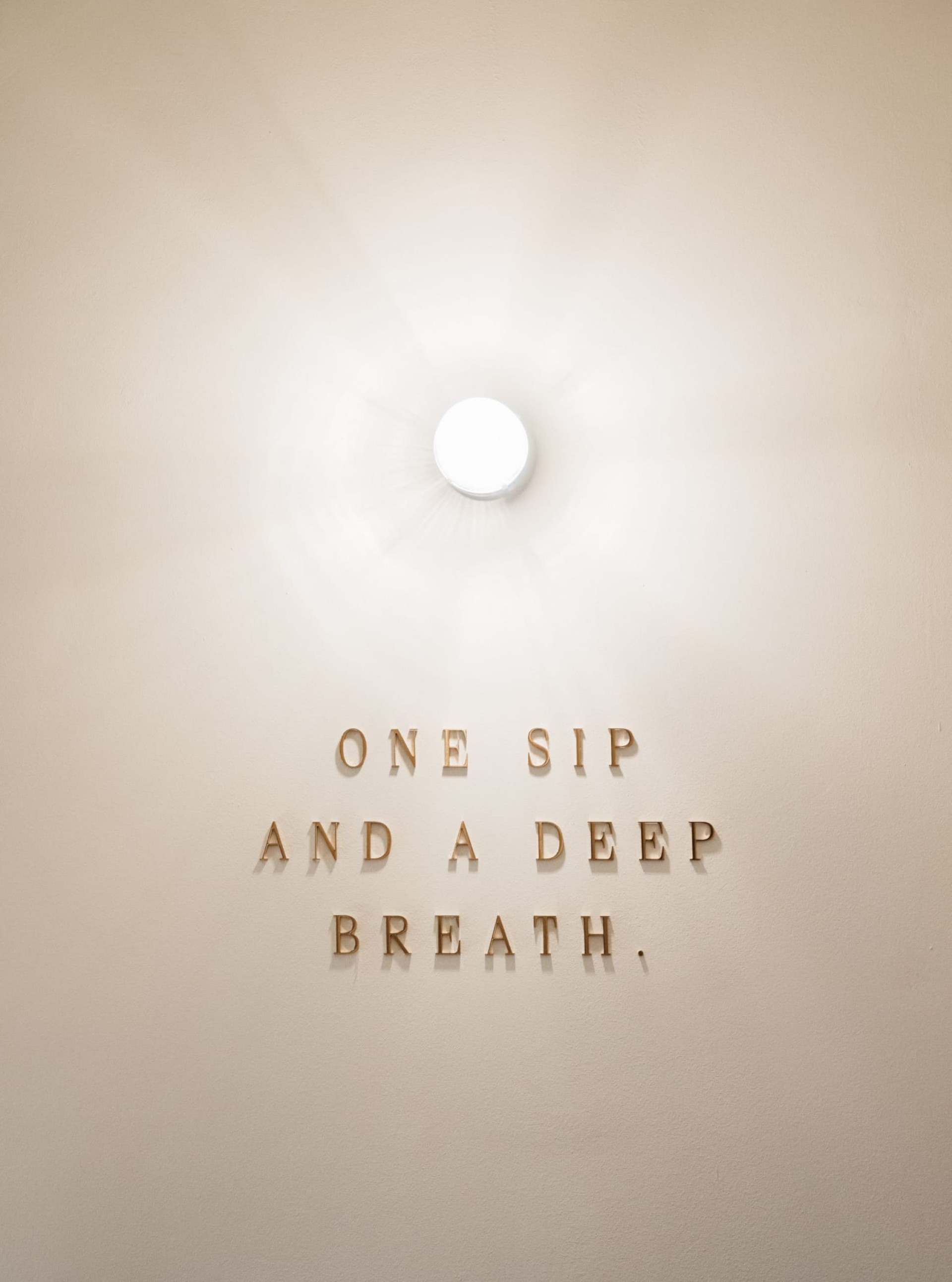 Beige Wand mit Lampe. Der Text lautet: "ONE SIP AND A DEEP BREATH."