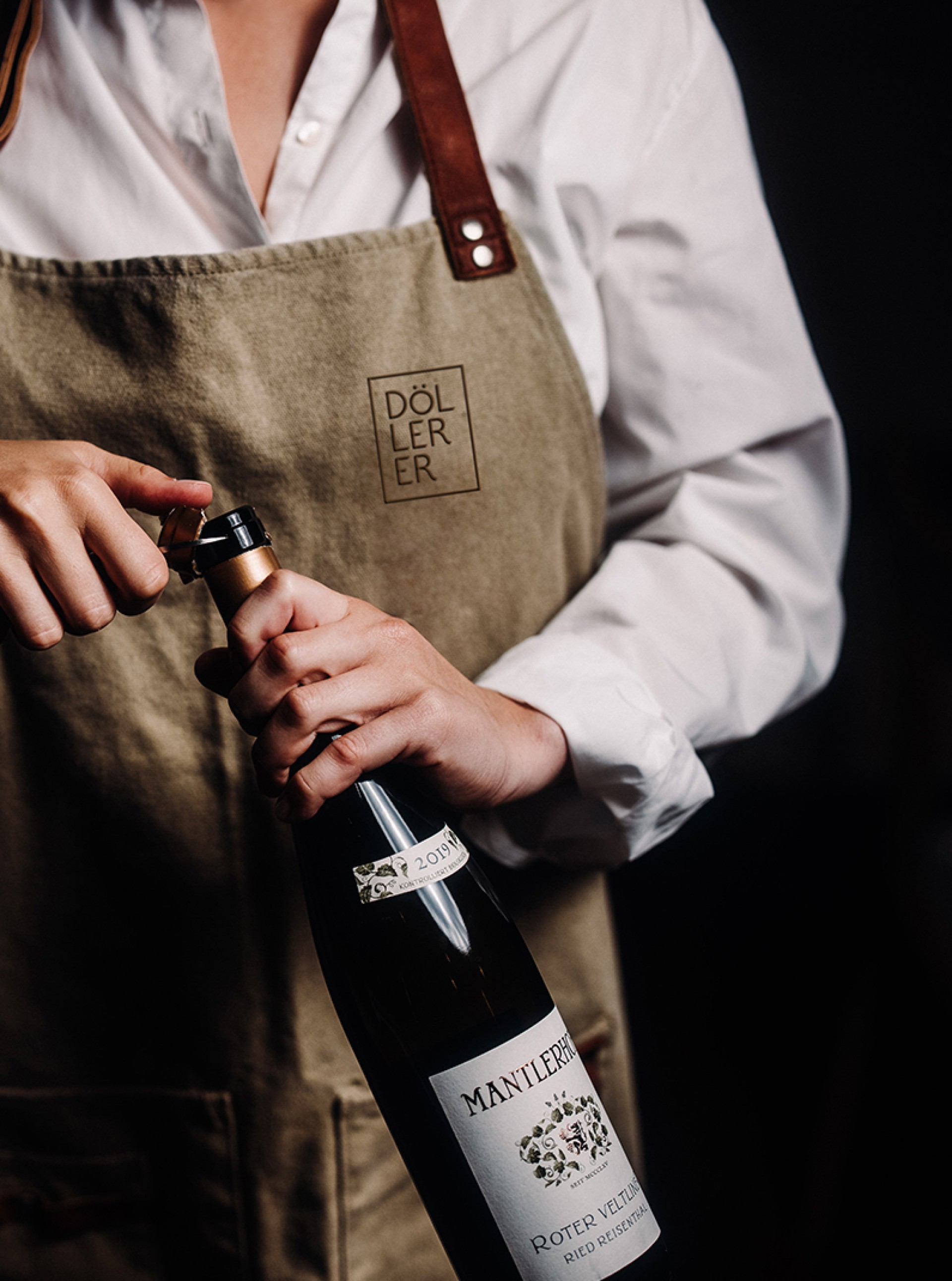 Eine Person öffnet eine Flasche "Mantlerhof Roter Veltliner Ried Reisenberg" mit "Döllerer" Schürze.