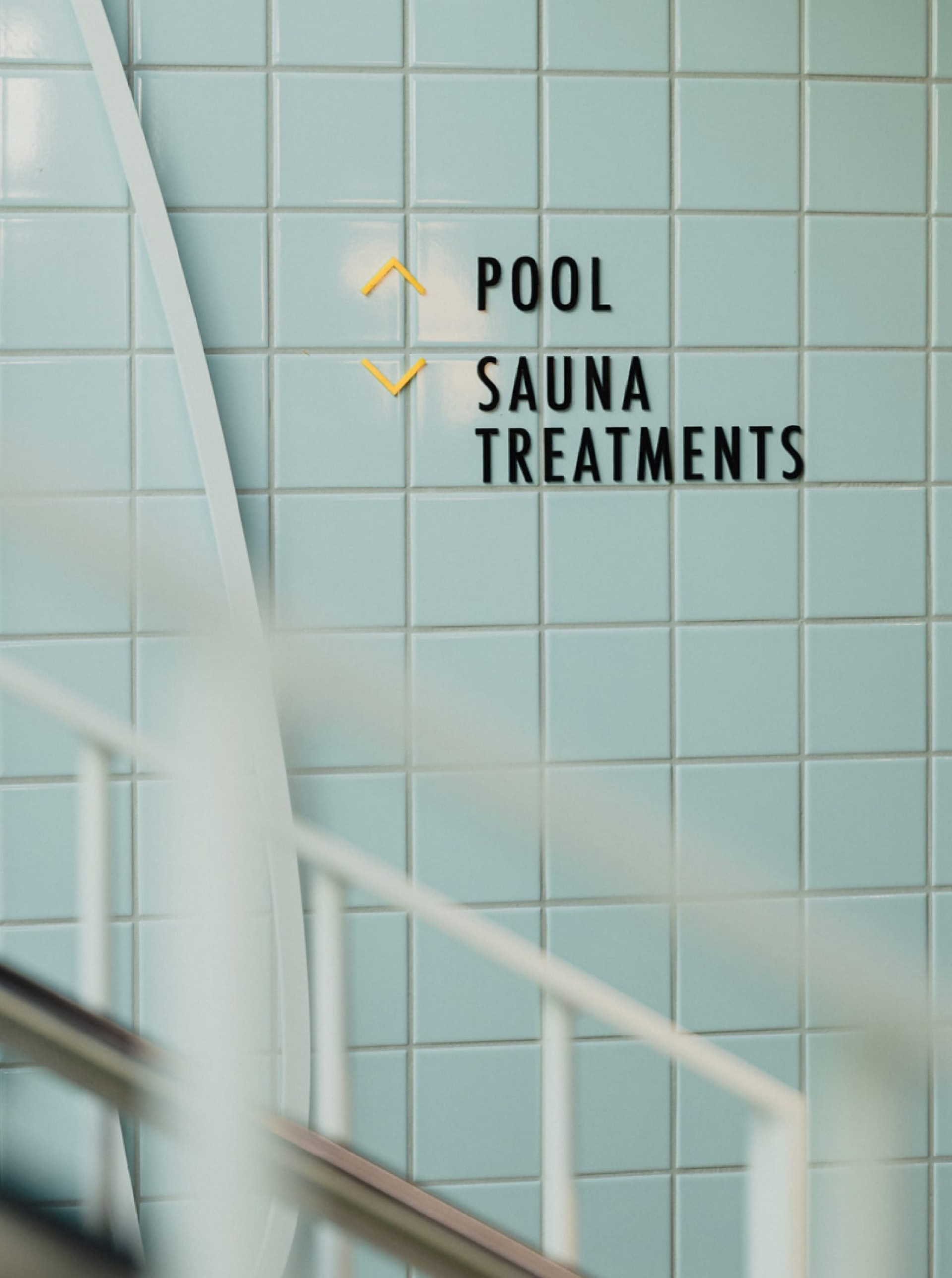 Ein Schild mit der Aufschrift "POOL", "SAUNA" und "BEHANDLUNGEN" mit Pfeilen auf einem blauen Fliesenhintergrund.
