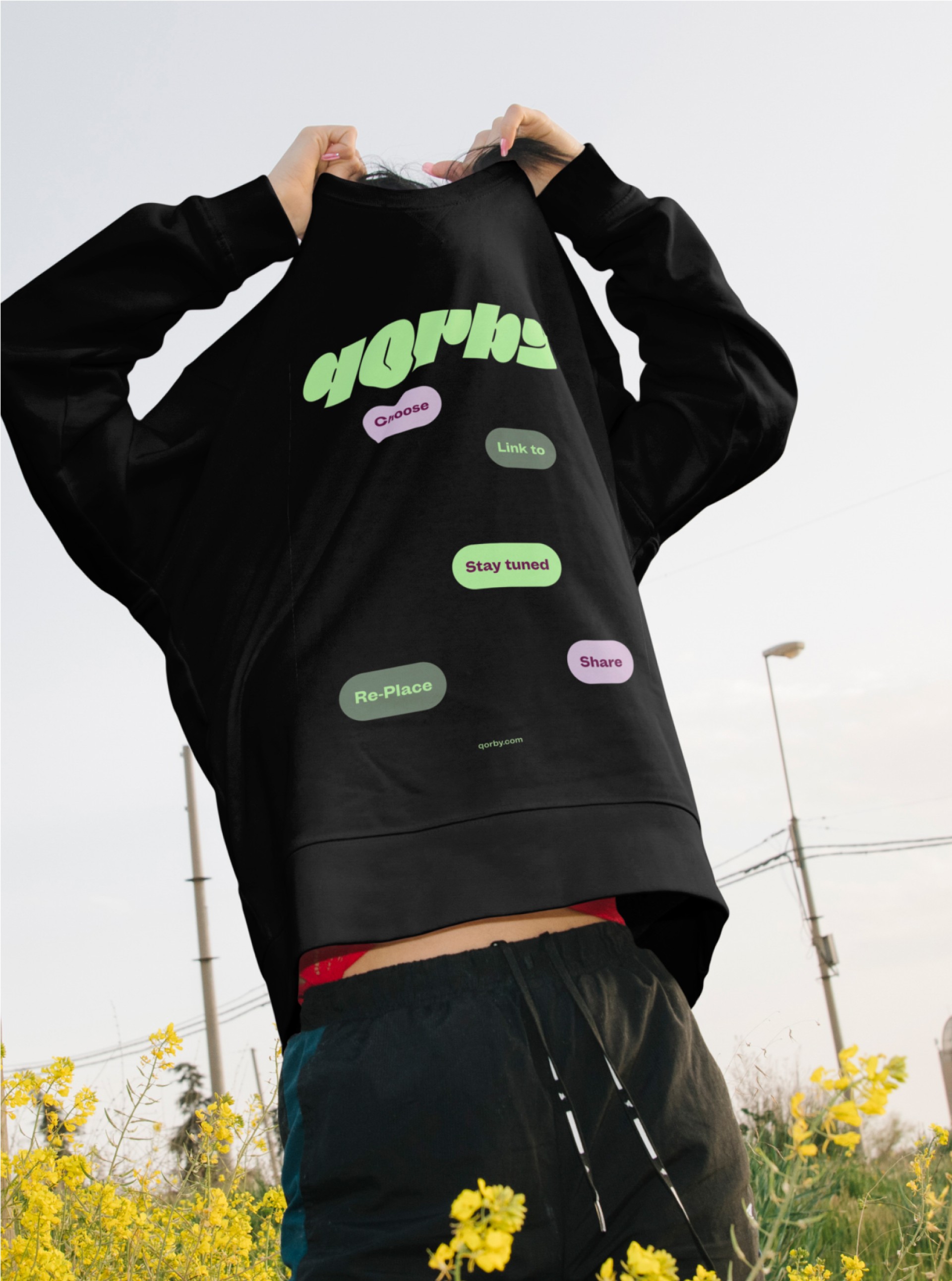 Person hält einen schwarzen "Works"-Pullover mit Aufdruck über dem Kopf in einem gelben Blumenfeld.