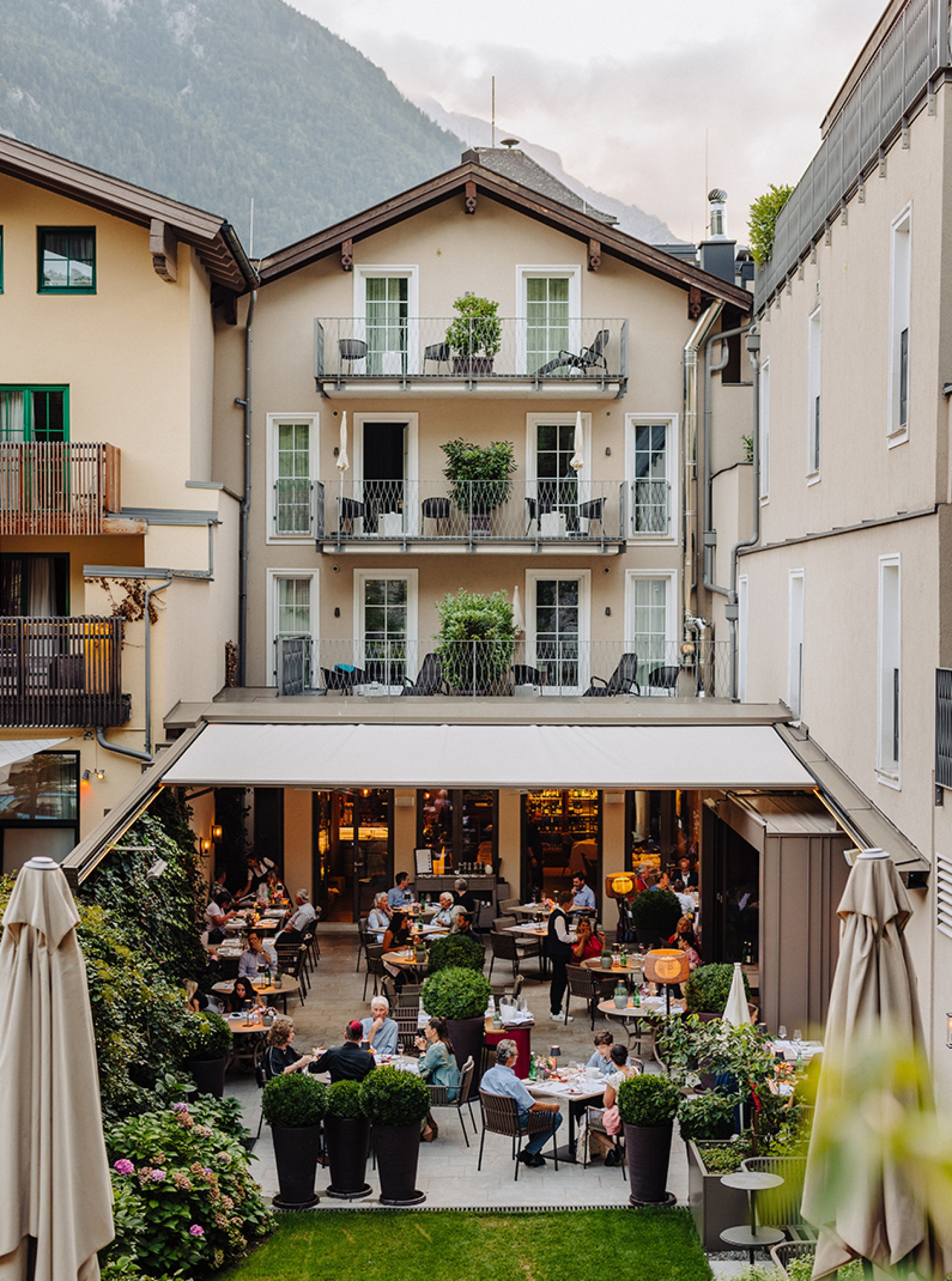 Außenansicht eines Restaurants mit Gästen auf der Terrasse, umgeben von Gebäuden und Bergen.