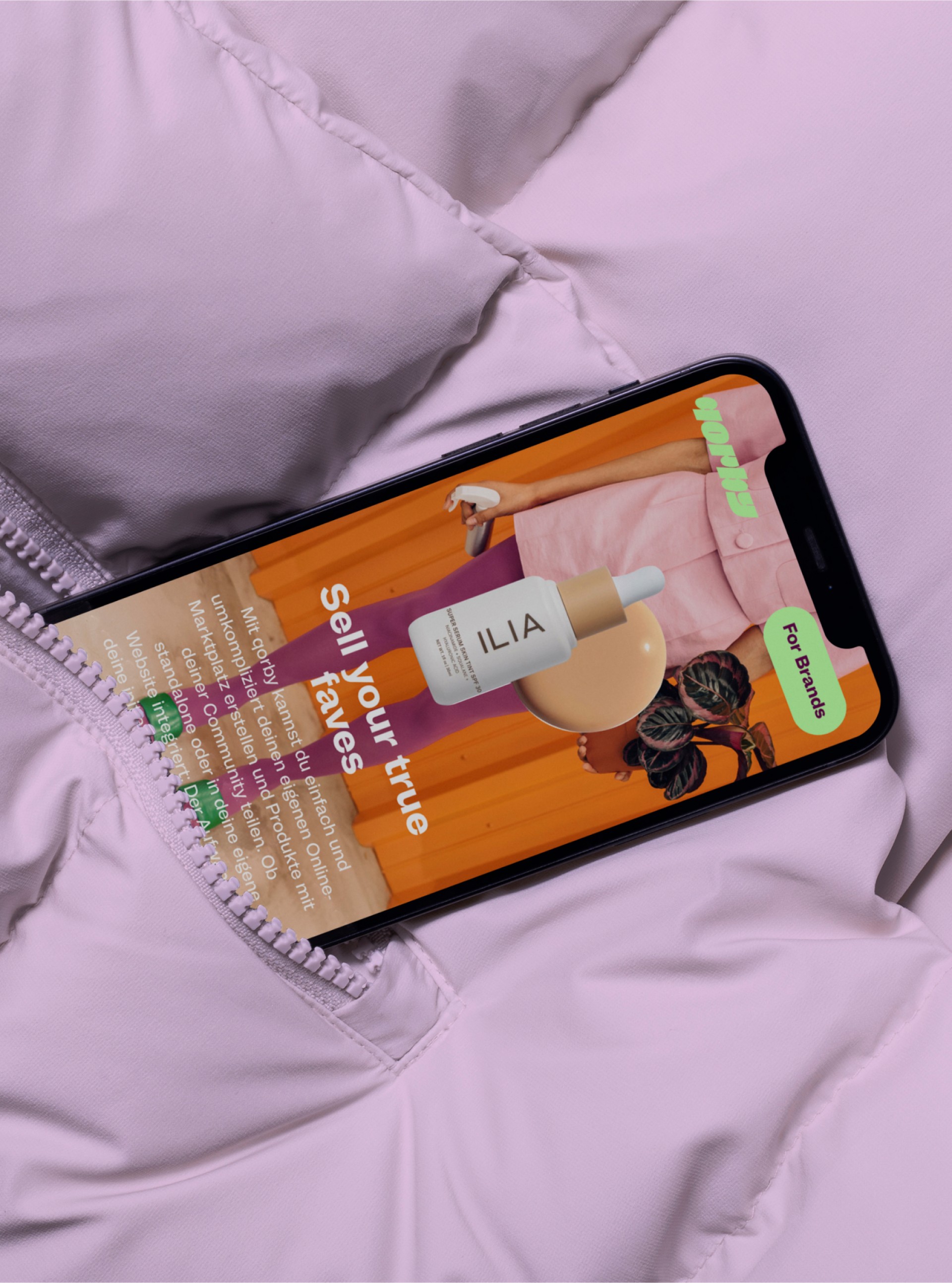 Ein Handy zeigt eine corby-Anzeige mit Ilia-Produkten auf einer rosafarbenen, gesteppten Jacke. Text: "Sell your true faves" & "For Brands".