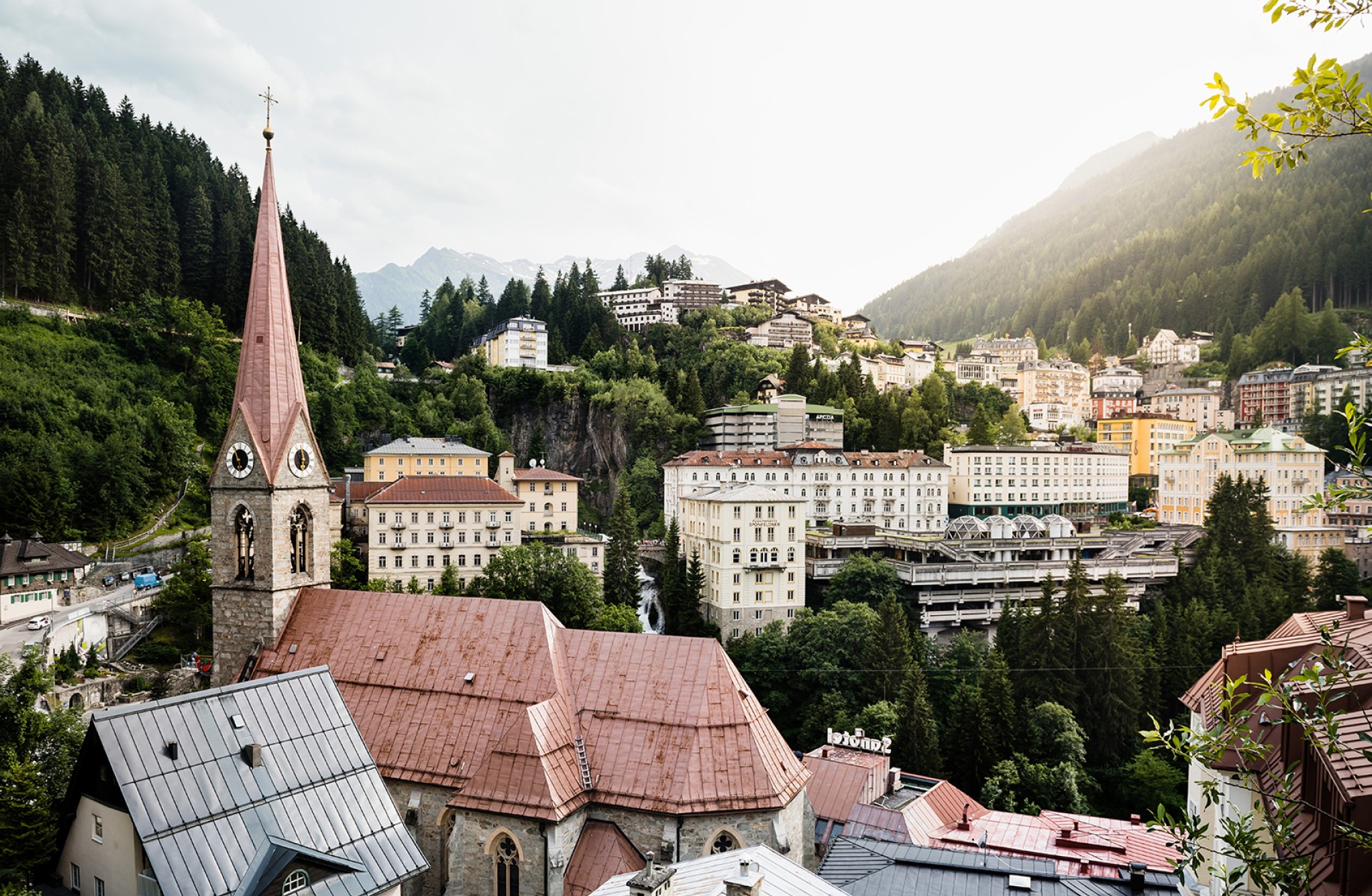 Ein Blick auf Bad Gastein, Österreich, mit einer Kirchturmspitze im Vordergrund, Gebäuden und bewaldeten Hügeln im Hintergrund.