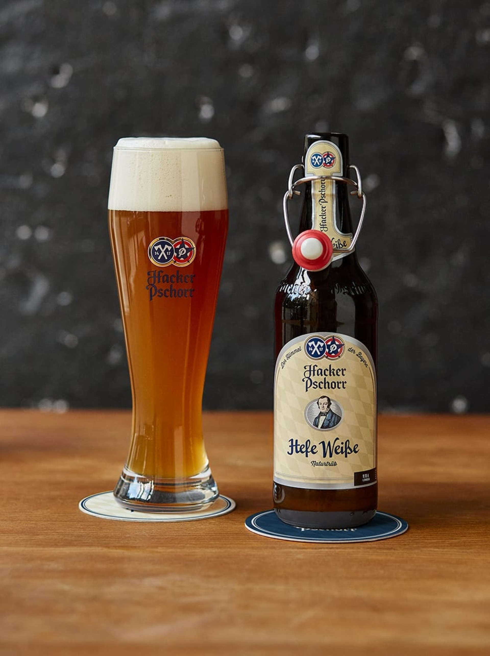 Ein Glas Hacker Pschorr Bier mit Schaum neben einer Flasche Hacker Pschorr Hefeweizen auf Holz.