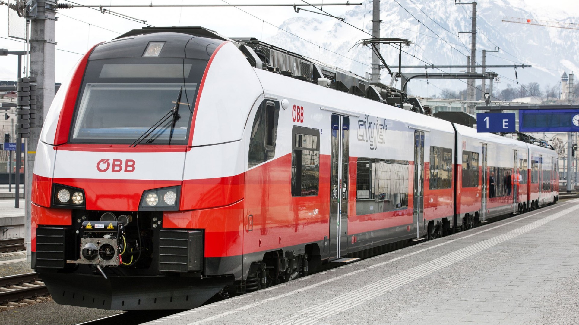Ein rot-weißer ÖBB Cityjet Zug steht an Bahnsteig 1E mit Bergen im Hintergrund.
