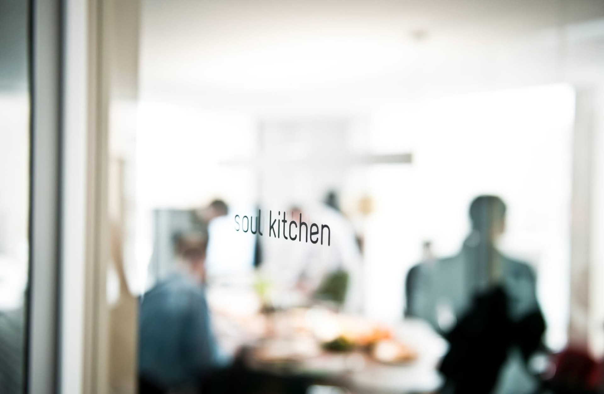 Verschwommene Ansicht durch Glastür mit Aufschrift "Soul Kitchen", Menschen um Tisch.