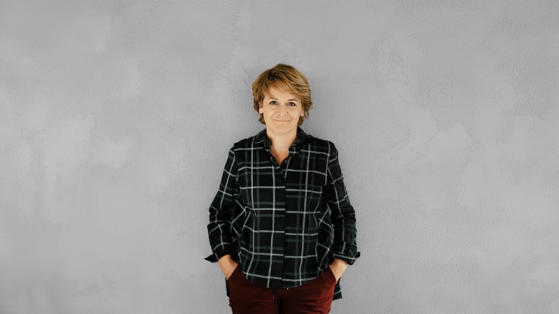 Eine Frau mit braunen Haaren, in kariertem Hemd und roten Hosen, steht vor einer grauen Wand.