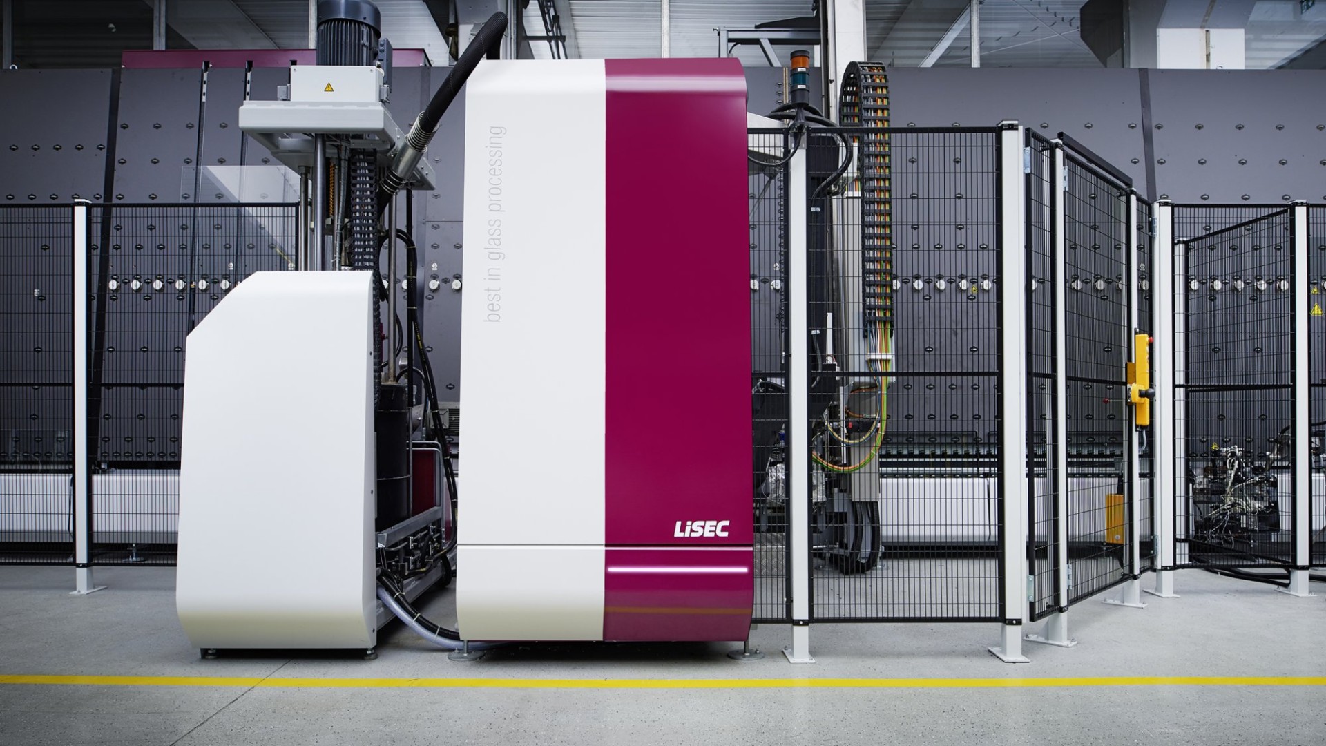 Eine LiSEC-Glasverarbeitungsmaschine, weiß und burgunderrot, steht hinter einem Maschendrahtzaun.