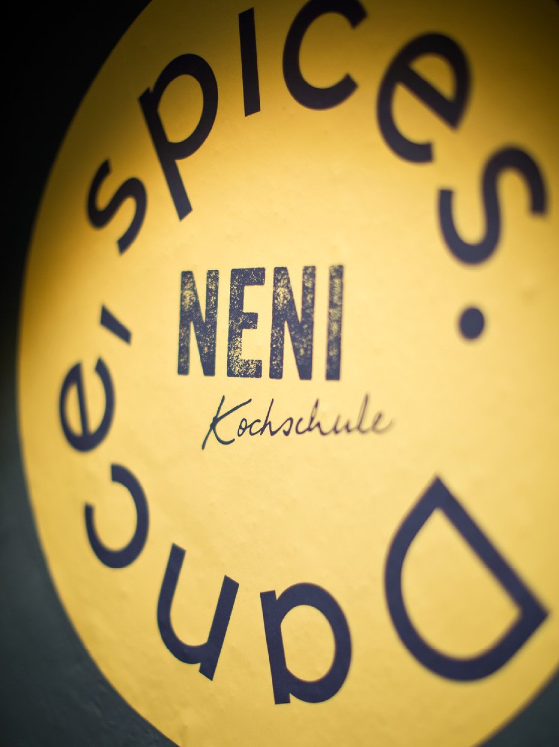 Gelbes NENI Kochschule Logo mit "spices & dance" in blauer Schrift darum.