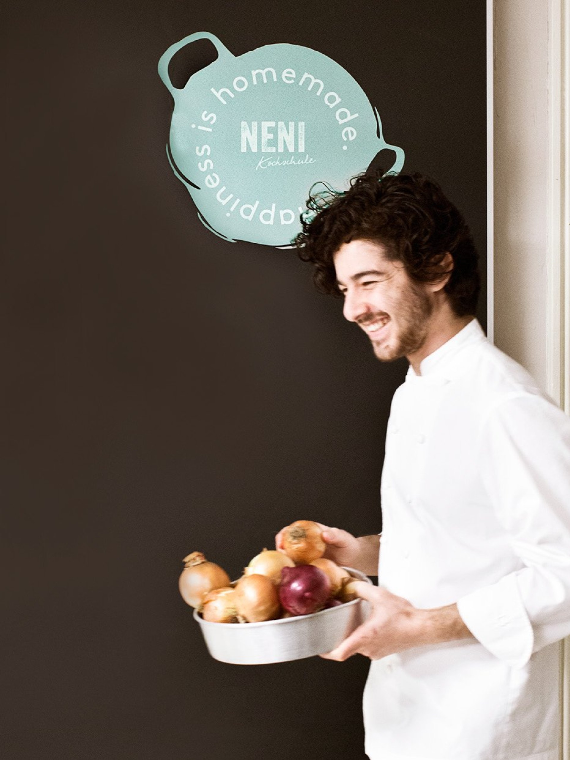 Ein lächelnder Koch mit Zwiebeln vor einer Tafel mit der Aufschrift "Happiness is homemade. Neni Kochschule".