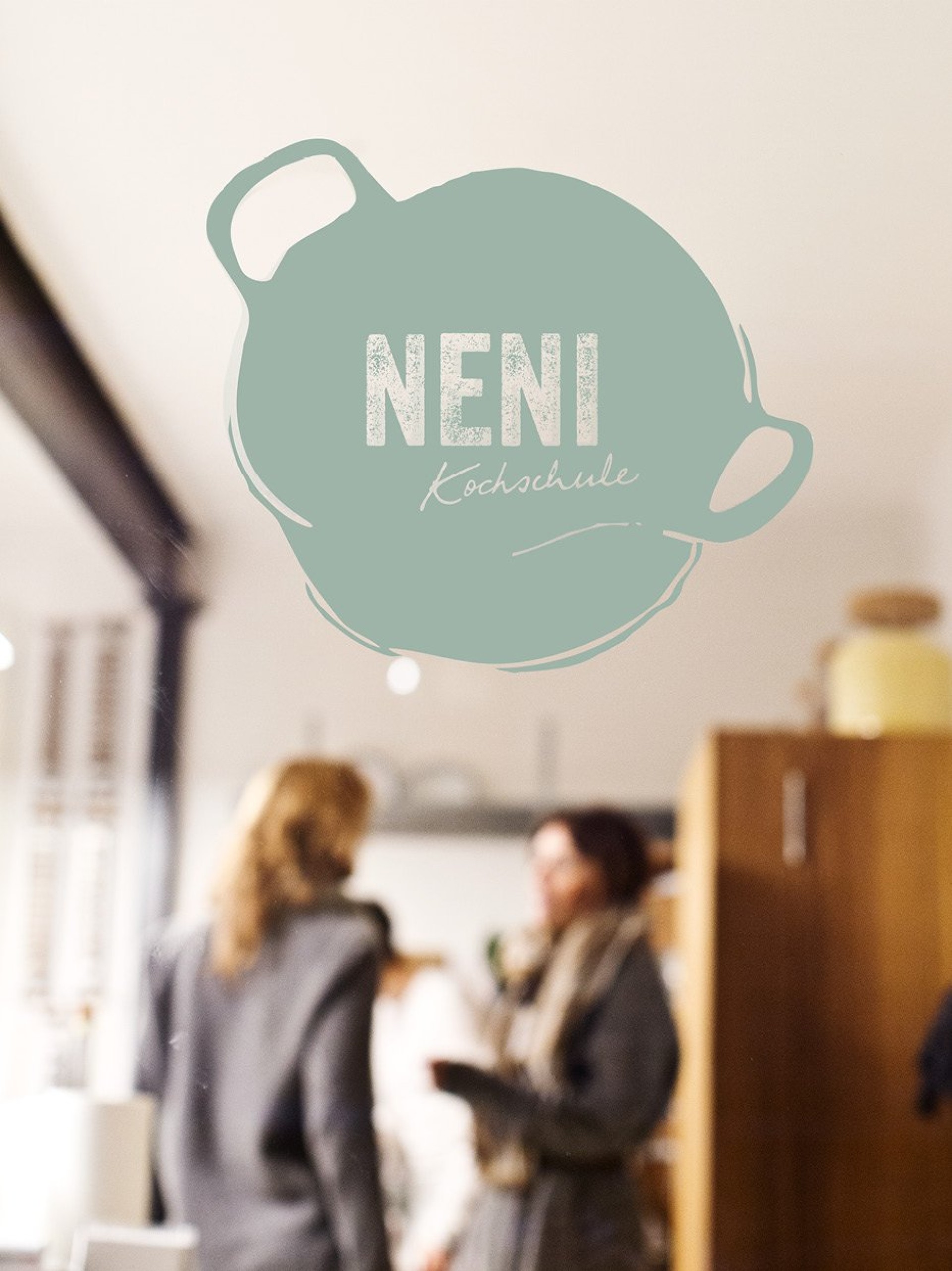 Logo von "Neni Kochschule" auf einer Glasscheibe mit zwei verschwommenen Personen im Hintergrund.