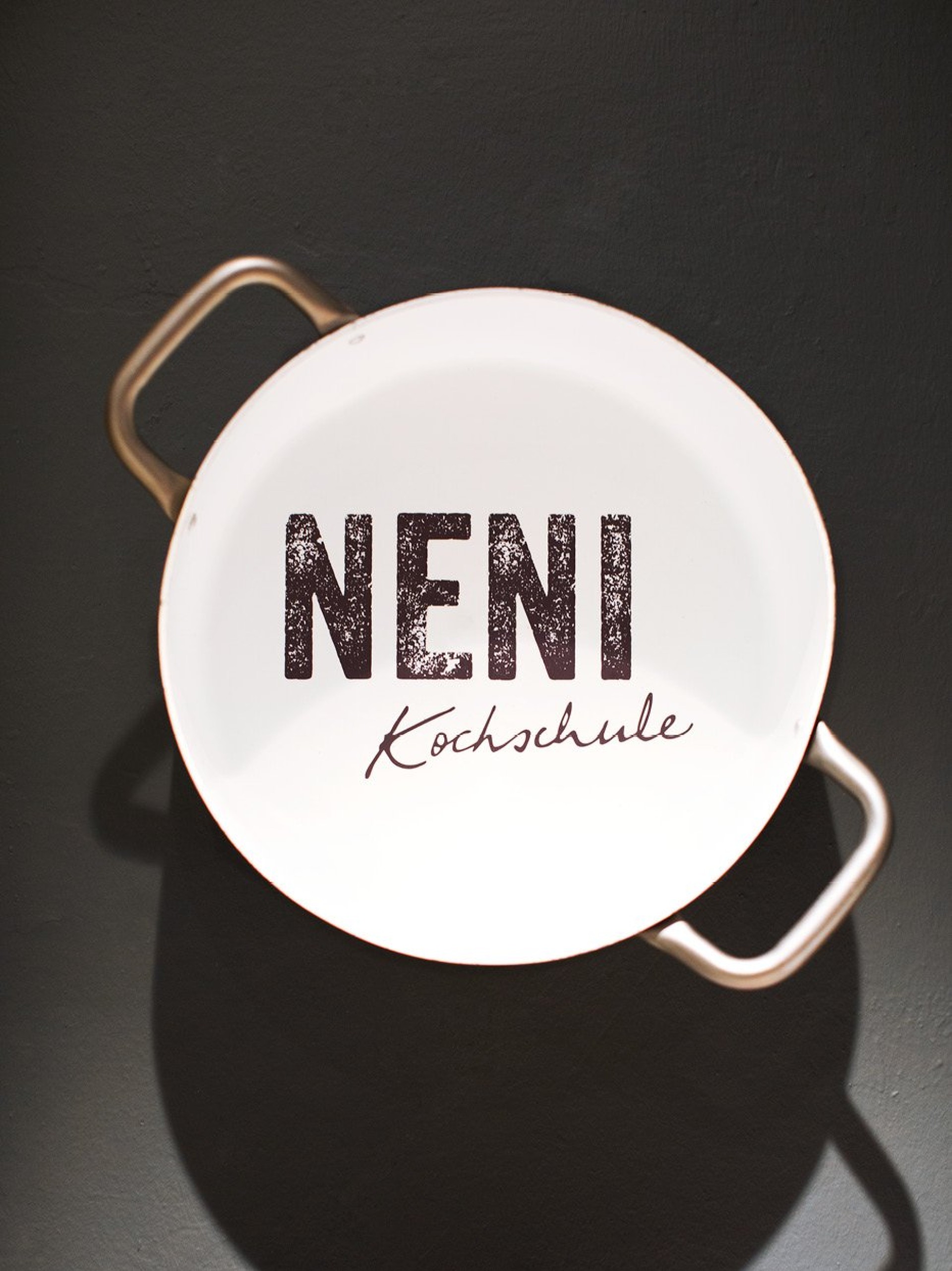 Weißer Kochtopf vor dunklem Hintergrund mit dem Text "NENI Kochschule" darauf.