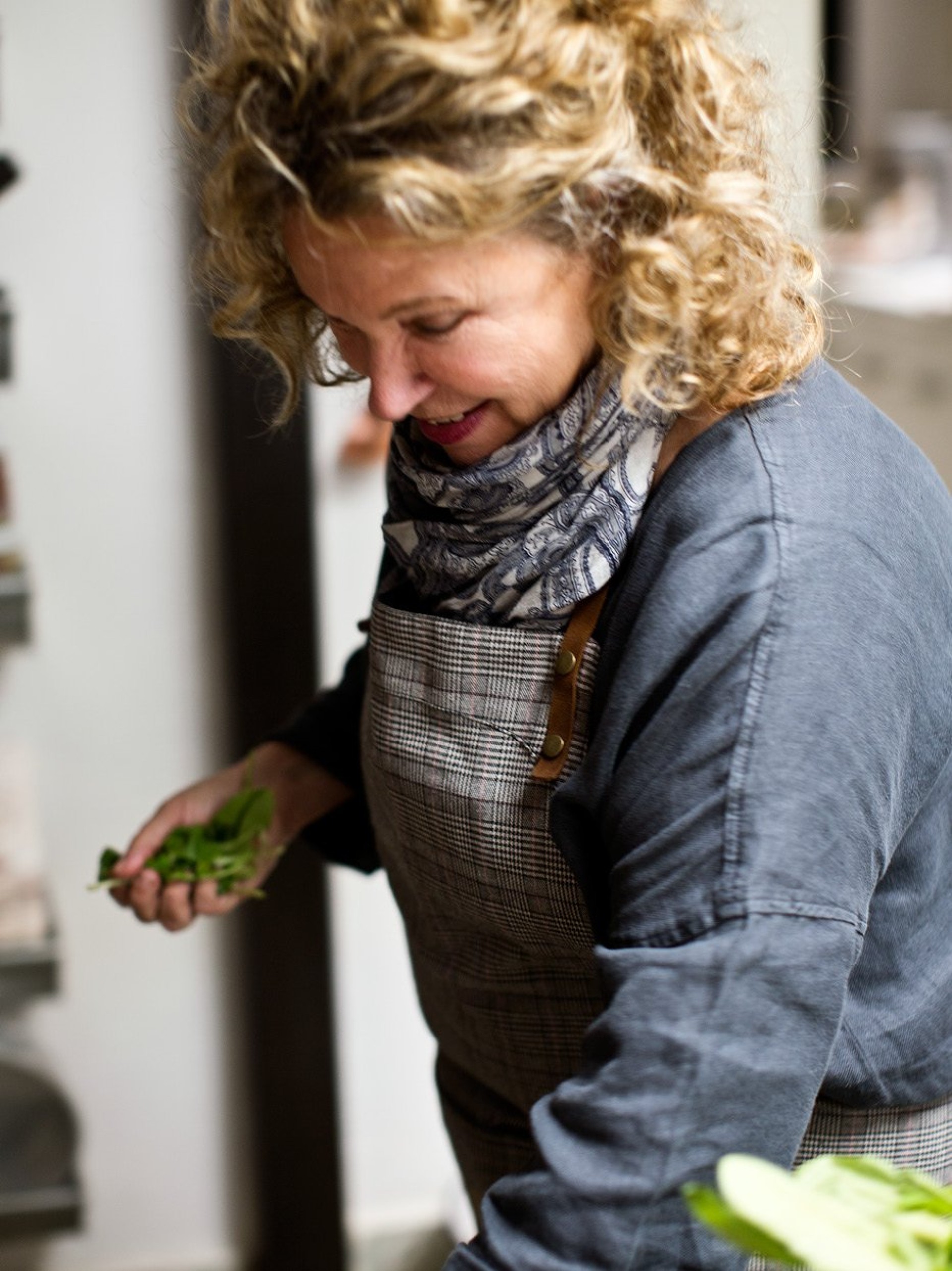 Eine Frau mit lockigem Haar betrachtet einen Haufen grüner Kräuter in der Hand. Sie trägt eine Schürze.