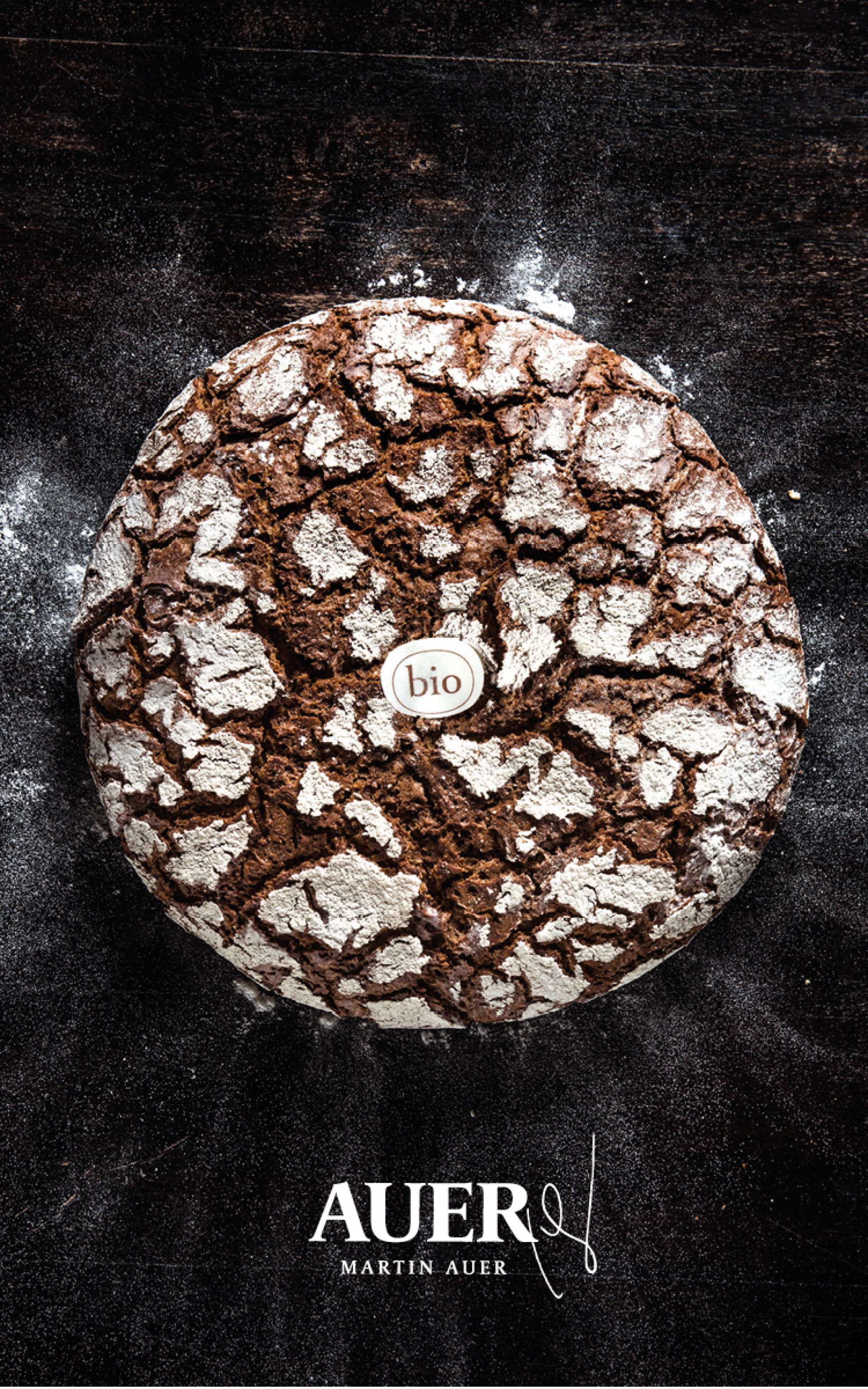 Ein Laib Bio-Brot von Martin Auer mit einem rissigen Äußeren auf einer dunklen Oberfläche mit AUER-Text darunter.