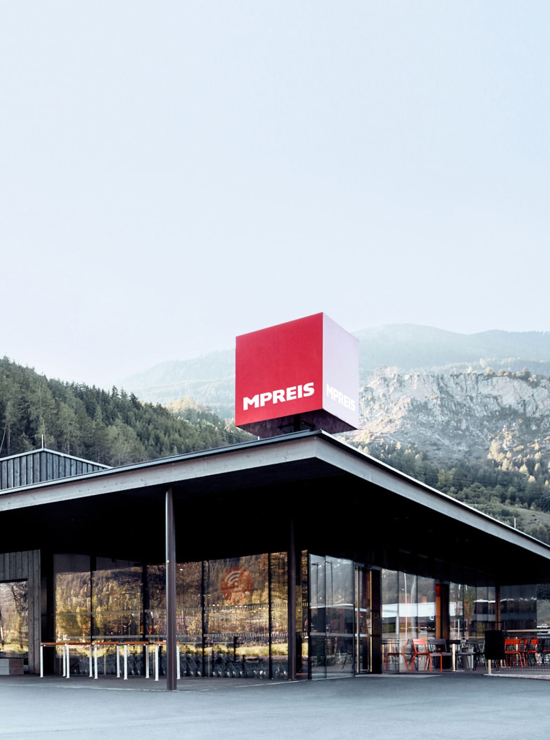 Ansicht eines Mpreis-Supermarkts mit rotem Logo auf einem Würfel vor bergiger Kulisse.