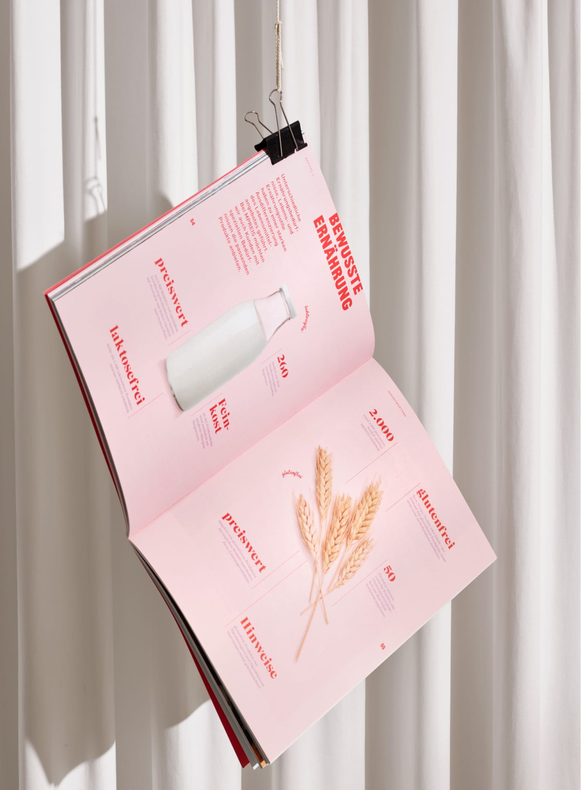 Geöffnetes Magazin "Bewusste Ernährung" mit rosa Seiten, Milch, Weizen, Text: preiswert, laktosefrei, glutenfrei. Vor weissen Vorhängen.