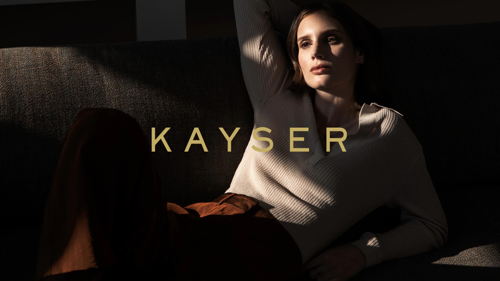 Frau liegt auf Sofa, trägt Pullover, Hose. Text „KAYSER“.