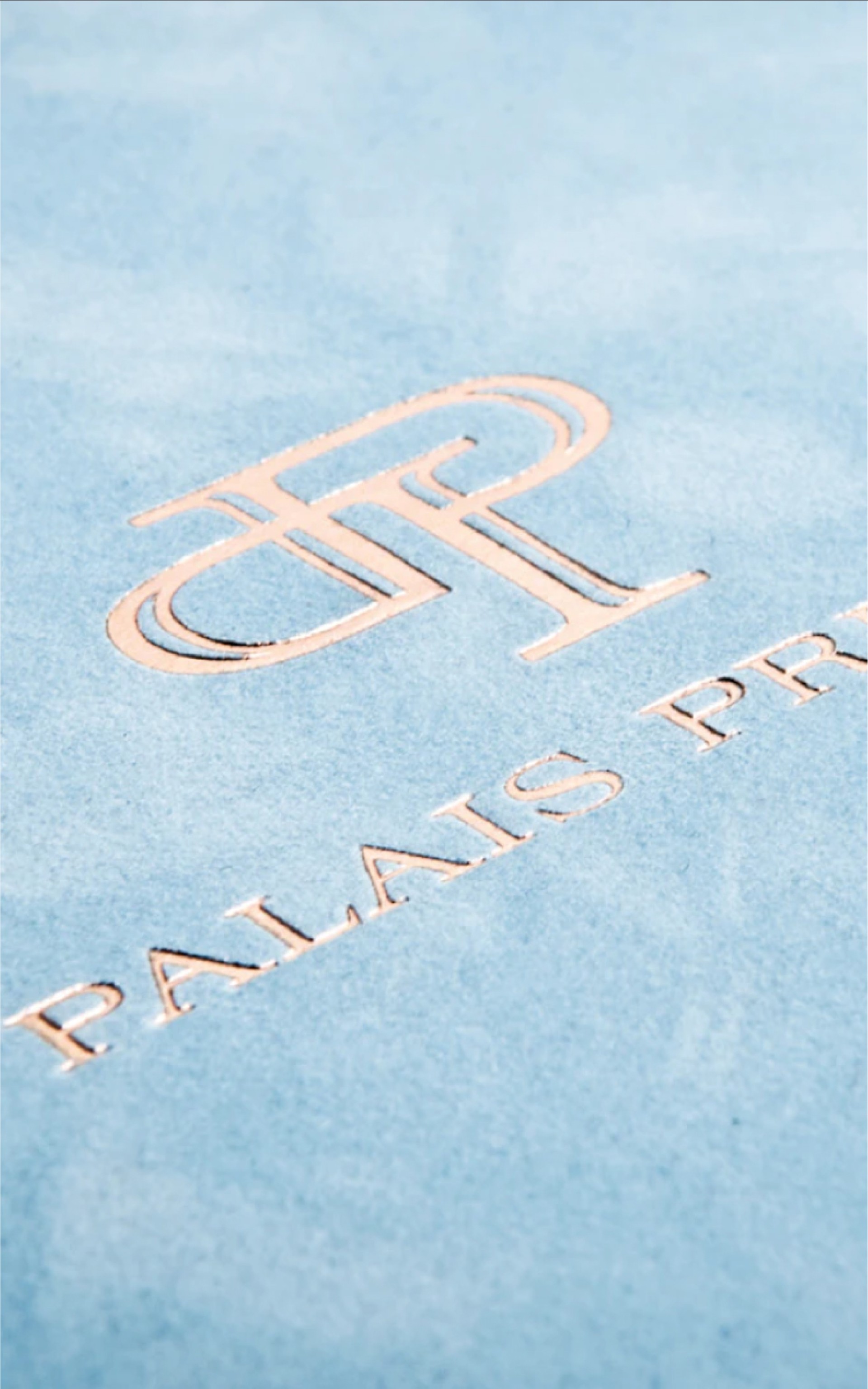Nahaufnahme des Logos und Textes "Palais Princier" in Roségold auf hellblauem Stoff.