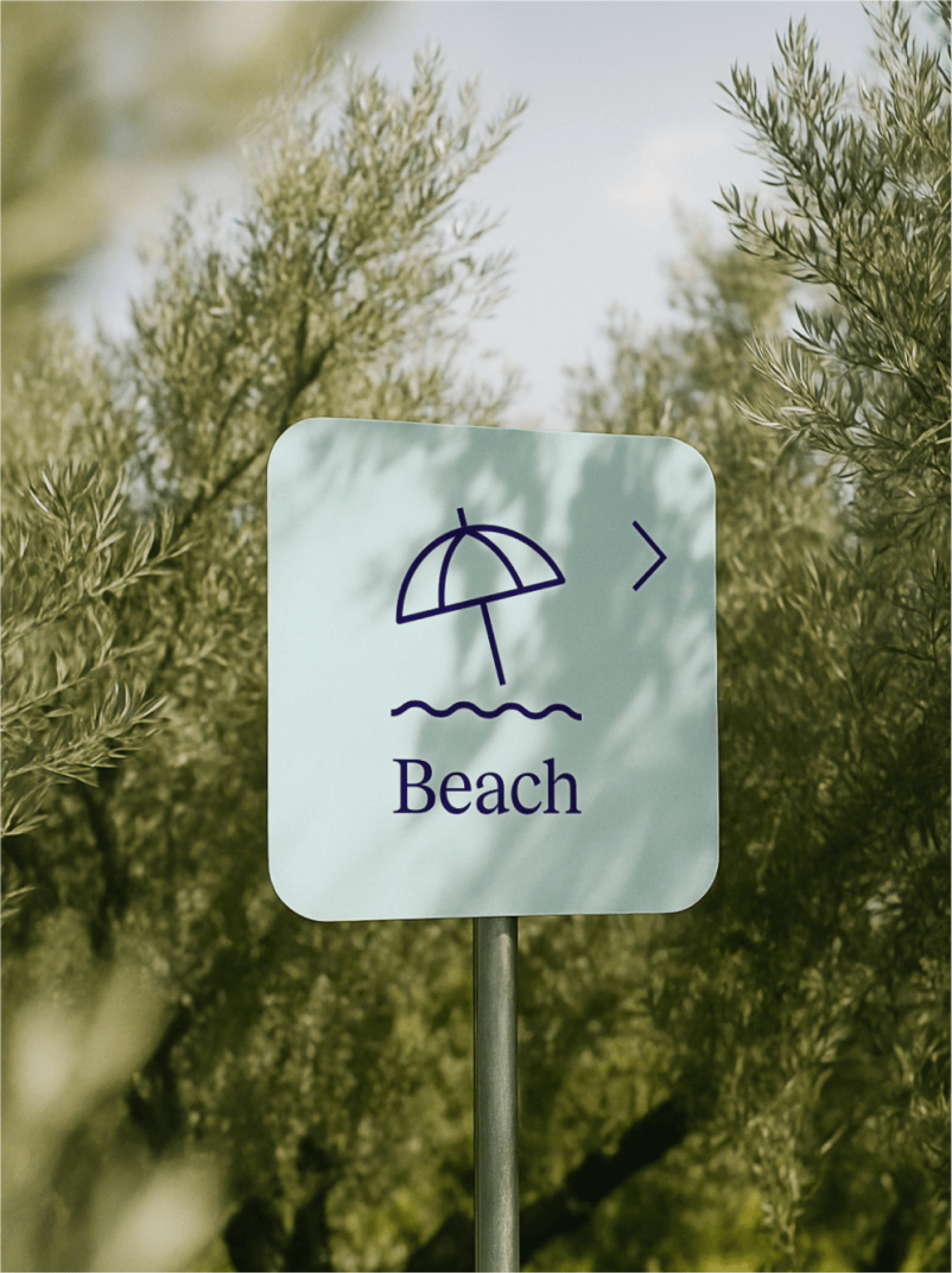 Ein hellblaues Schild mit der Aufschrift "Beach", einem Symbol für einen Sonnenschirm und einem Pfeil nach rechts, steht zwischen Bäumen.