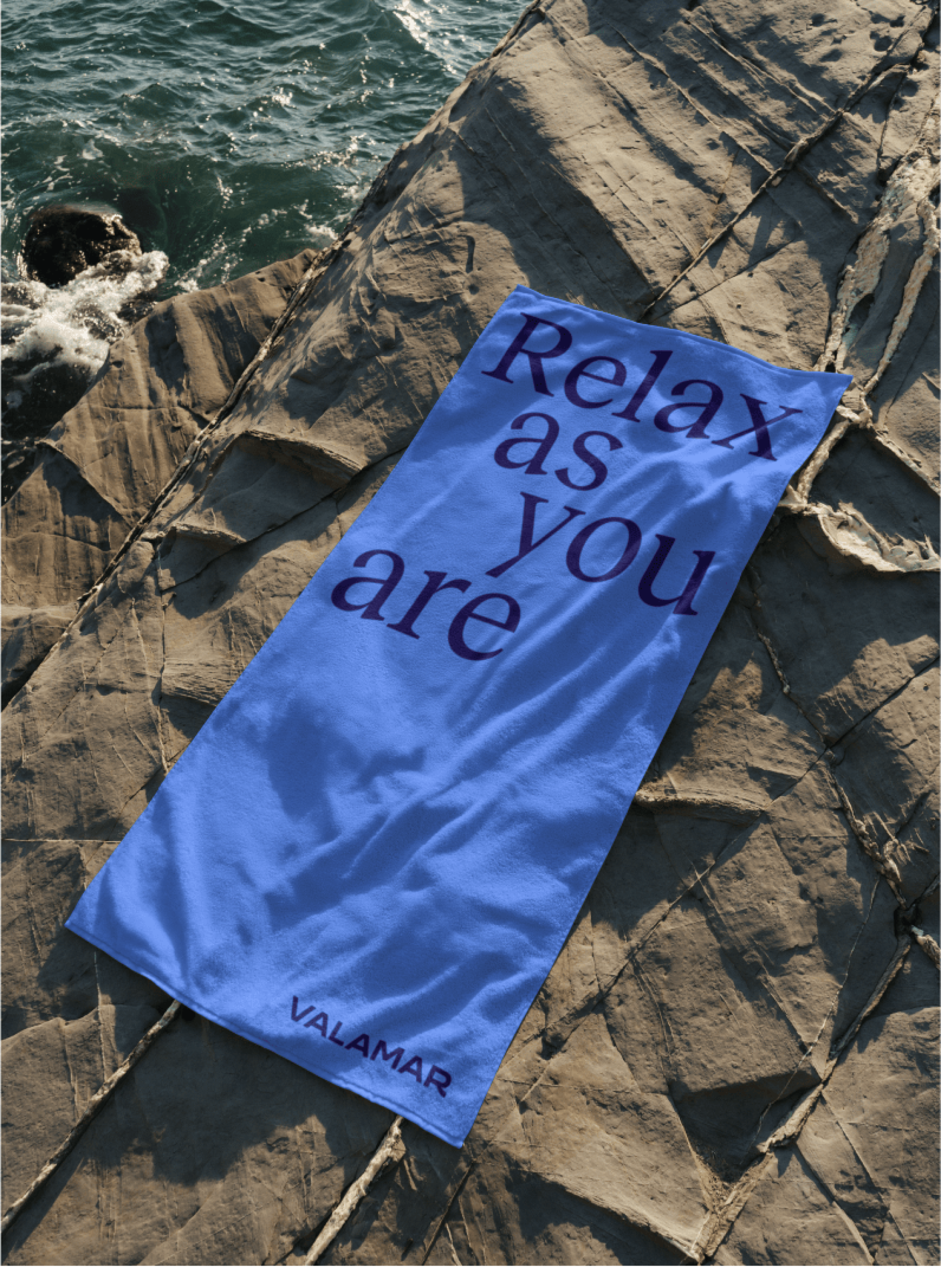 Ein blaues VALAMAR-Handtuch auf einem Felsen am Meer mit dem Text „Relax as you are“.