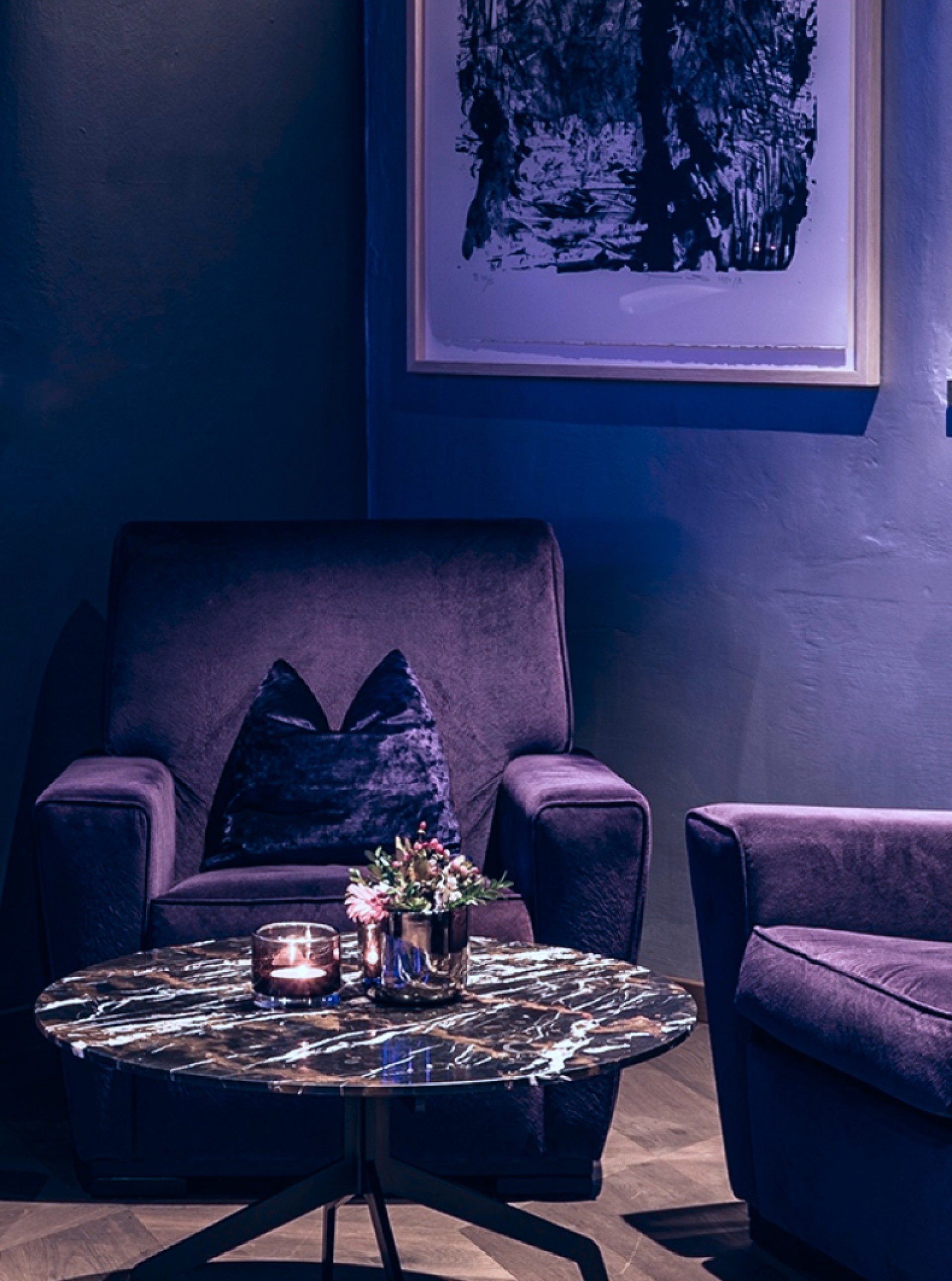 Lila Sessel, Couch und Marmortisch vor einer Wand mit Kunst. Kerze und Blumen drauf.
