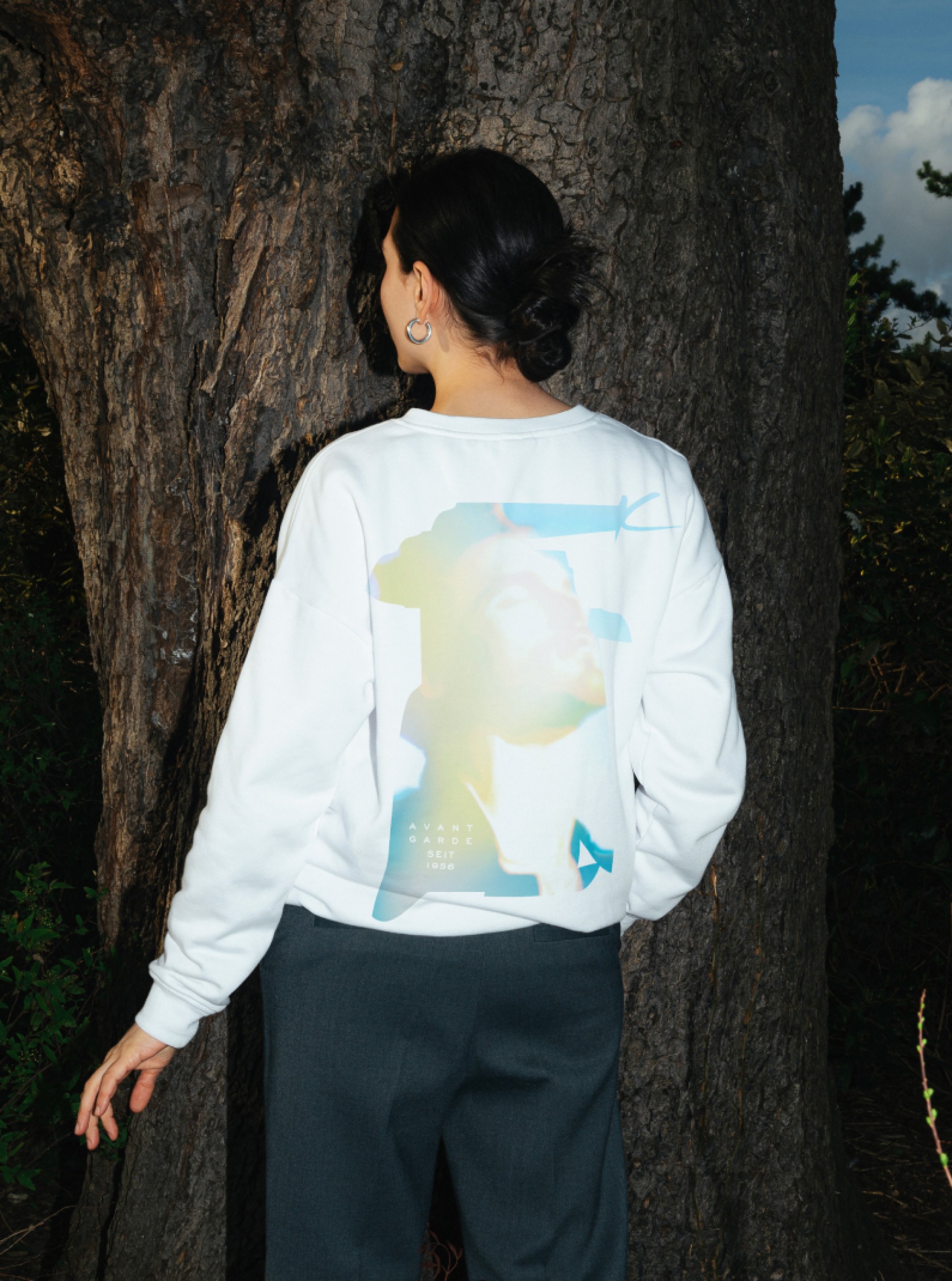 Frau mit Avant Garde Sweatshirt lehnt an Baum. Aufdruck mit Porträt und Text "Avant Garde Seit 1996".