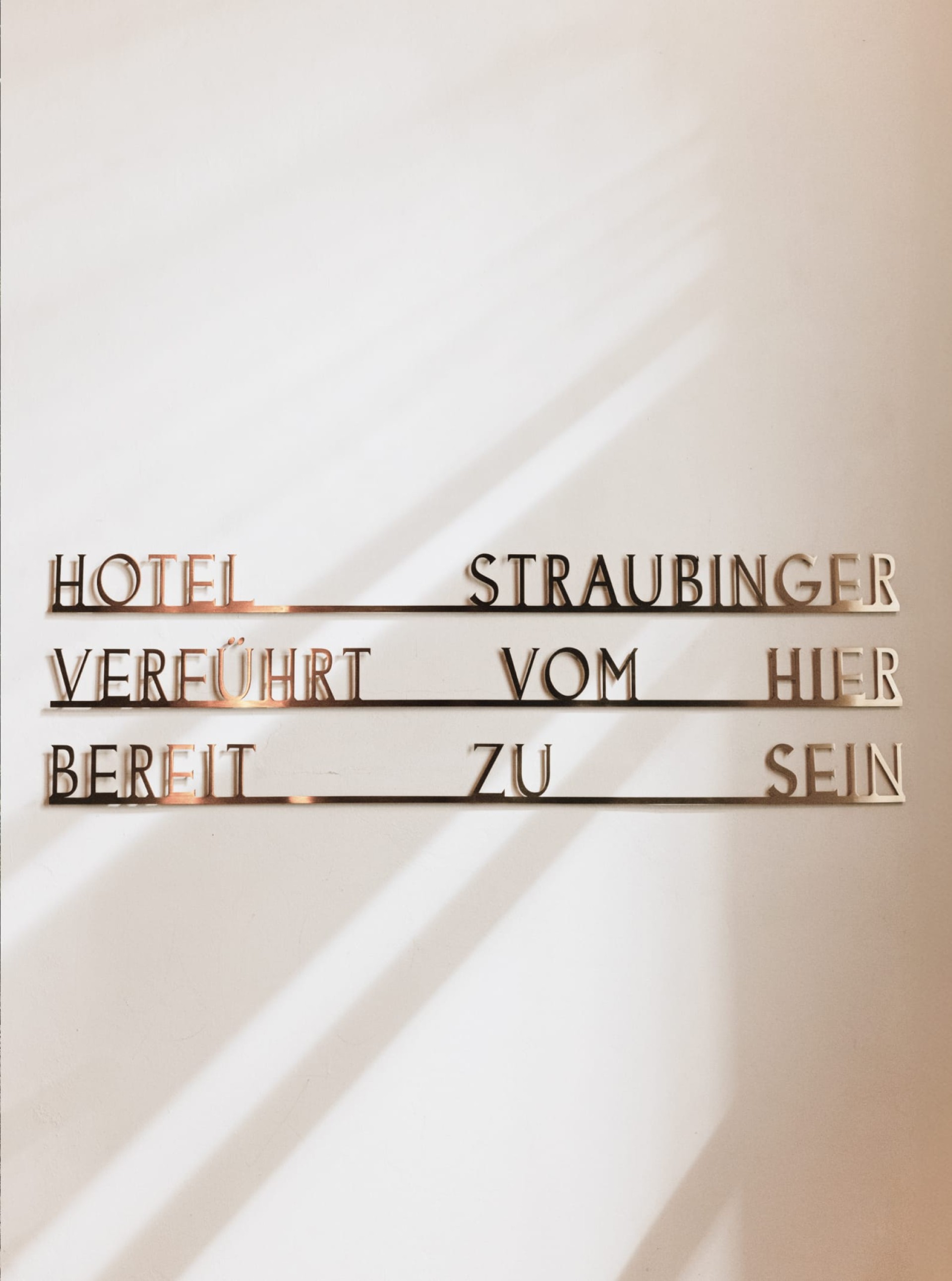 "Hotel Straubinger, verführt vom Hier, bereit zu sein" auf einer weißen Wand mit Schatten.