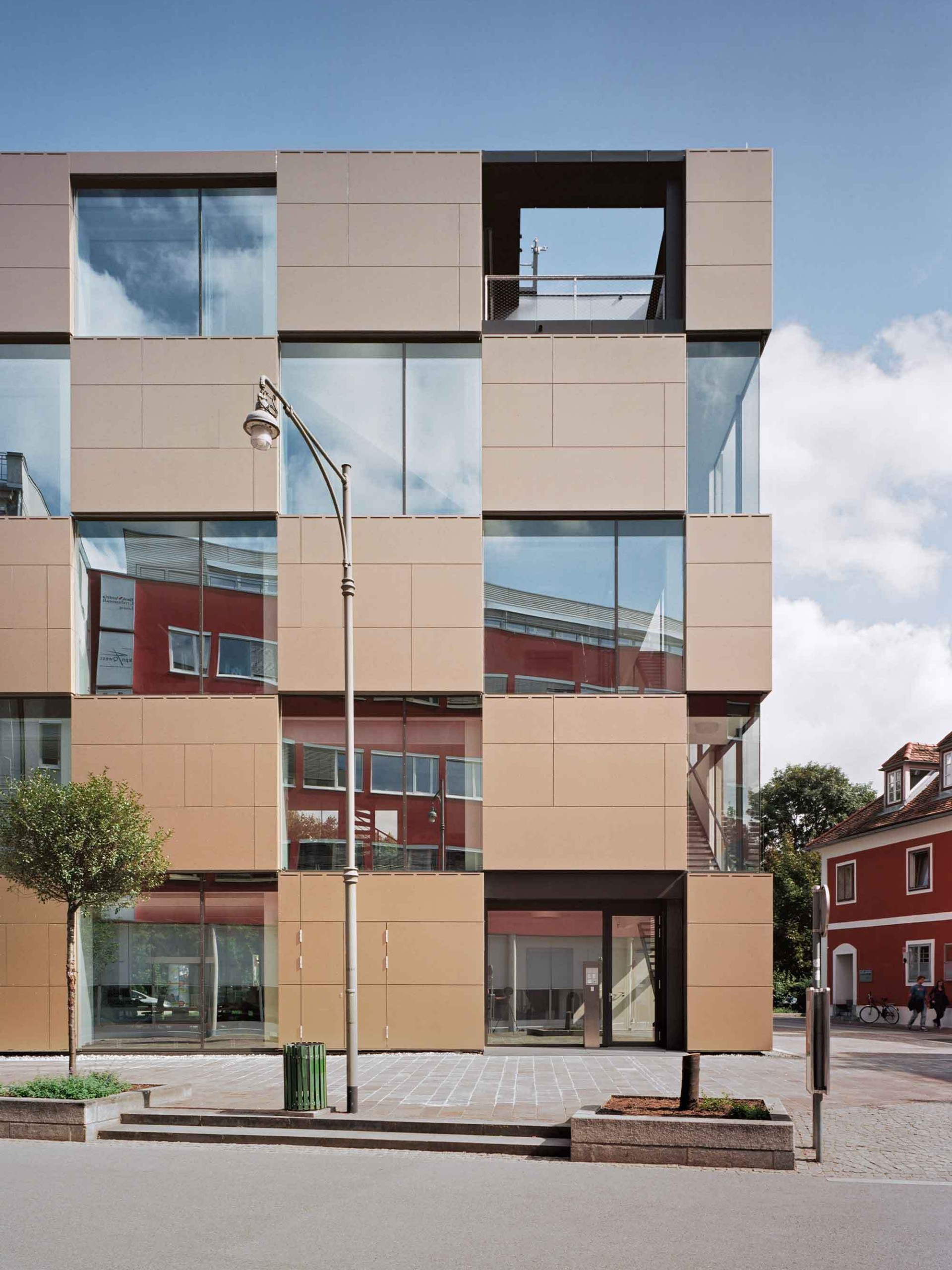 Ein Gebäude mit quadratischen Fenstern und braunen Paneelen. Spiegelungen von Himmel und Nachbargebäuden sind in den Fenstern sichtbar.