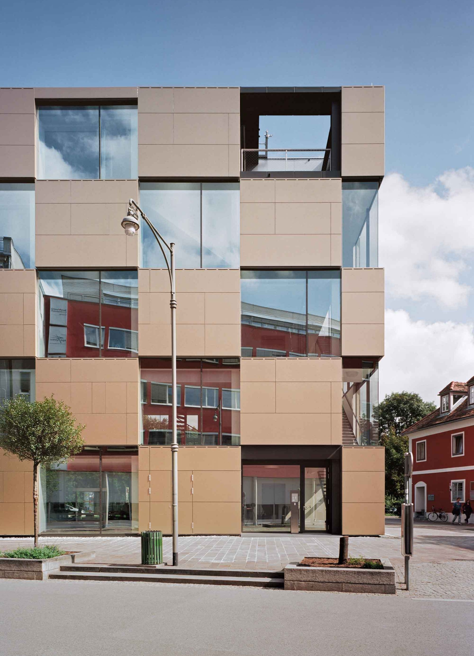 Ein Gebäude mit quadratischen Fenstern und braunen Paneelen. Spiegelungen von Himmel und Nachbargebäuden sind in den Fenstern sichtbar.