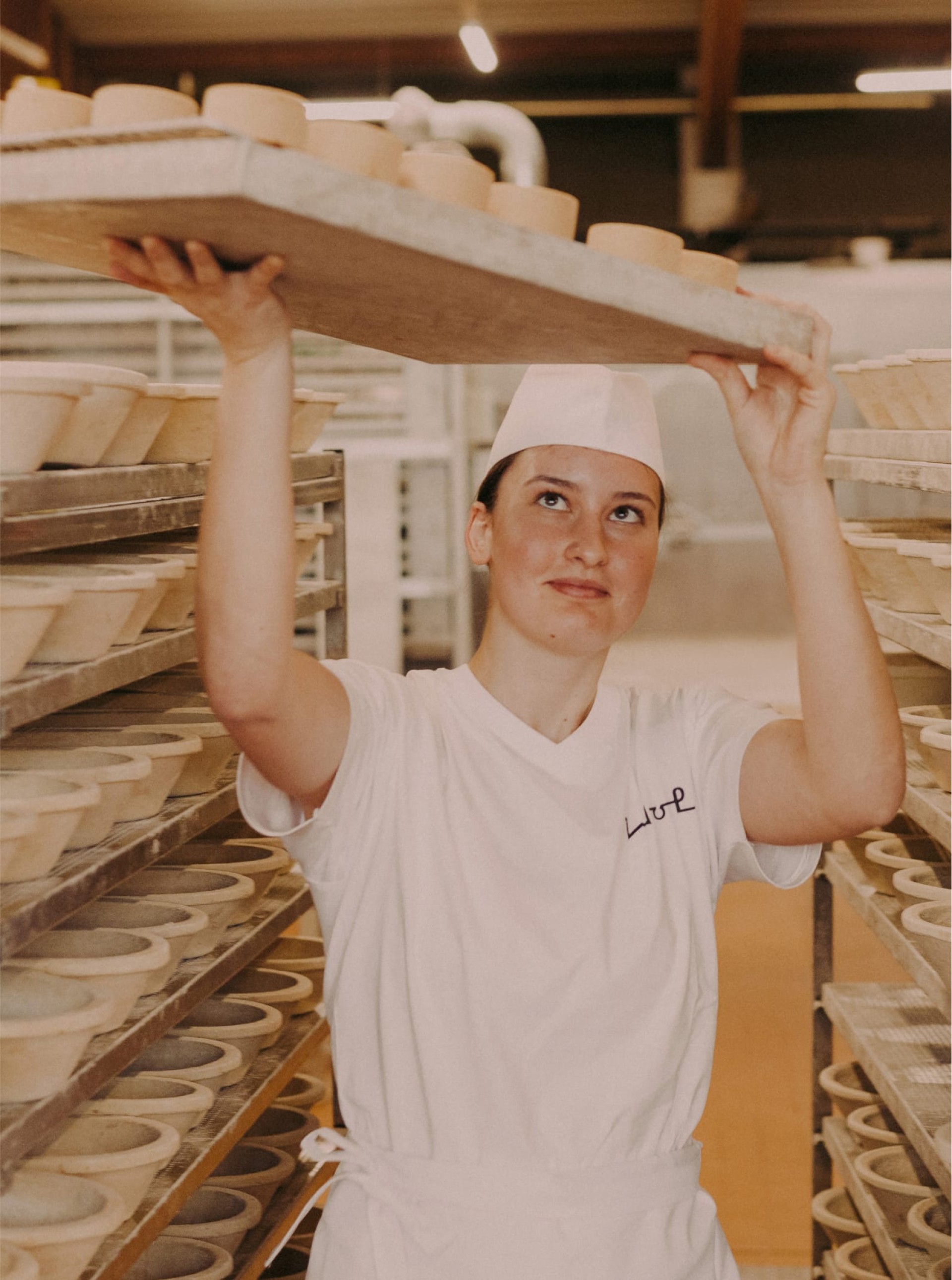 Eine Bäckerin, die ein Tablett mit Laiben über Regalen in einer Bäckerei hält.