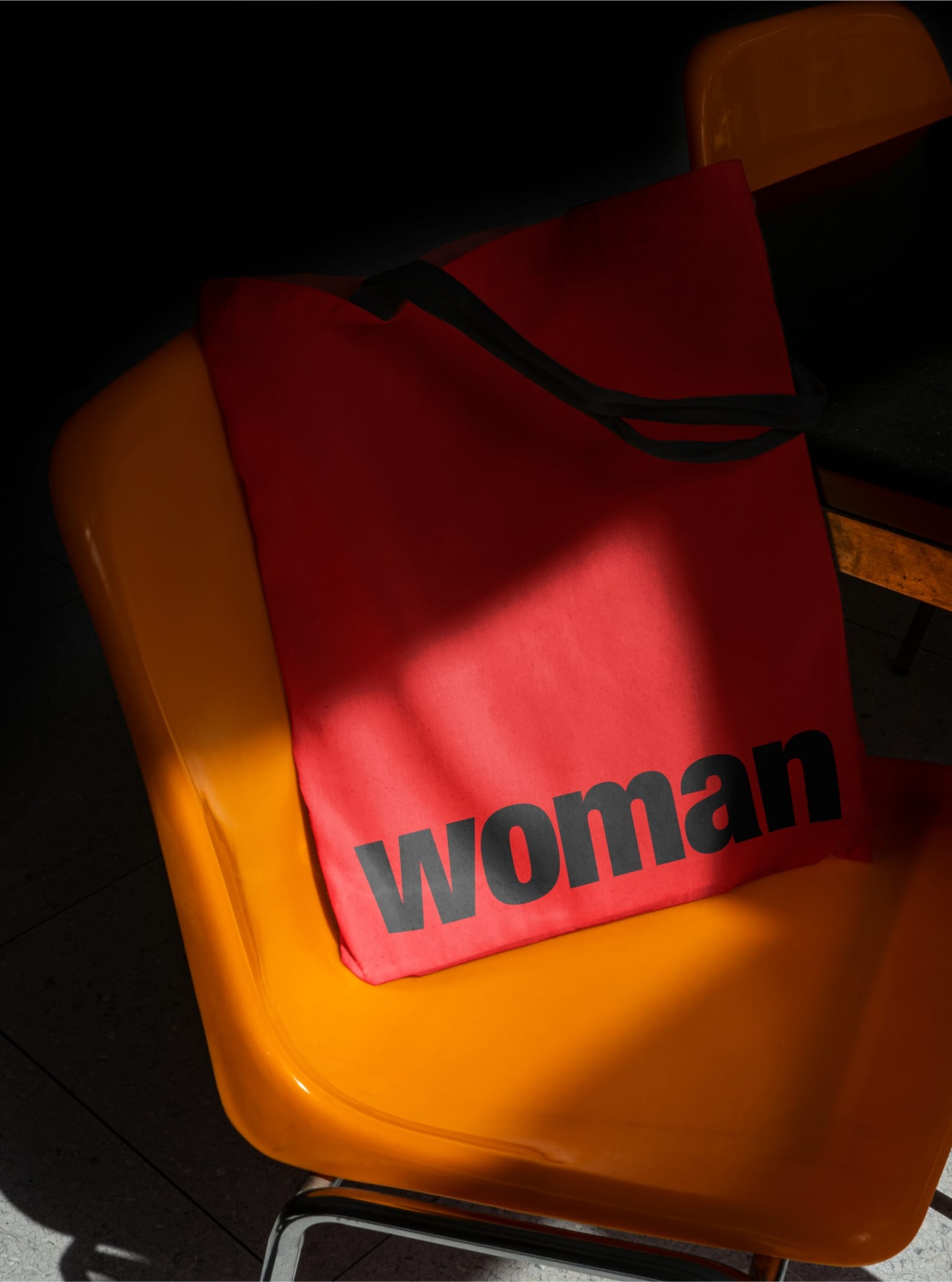 Eine rote Einkaufstasche mit der Aufschrift "Woman" liegt auf einem orangefarbenen Stuhl im Sonnenlicht.
