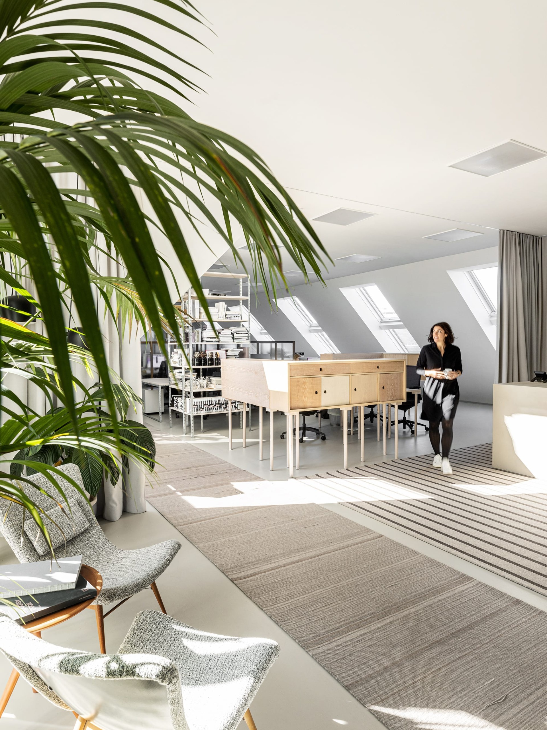 Eine Frau in Schwarz geht in einem hellen Büro mit Pflanzen, modernen Möbeln und Dachfenstern.