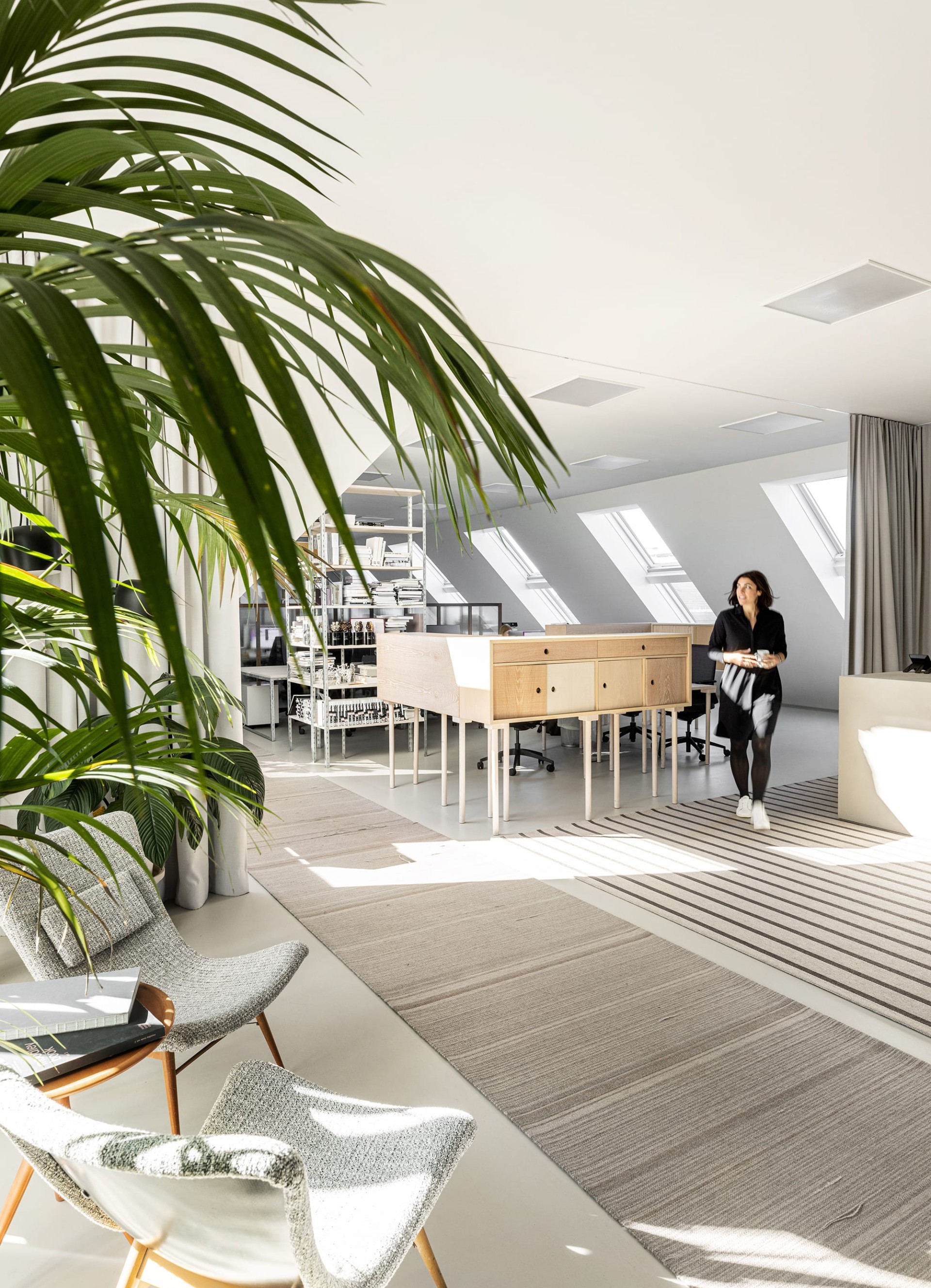 Eine Frau in Schwarz geht in einem hellen Büro mit Pflanzen, modernen Möbeln und Dachfenstern.