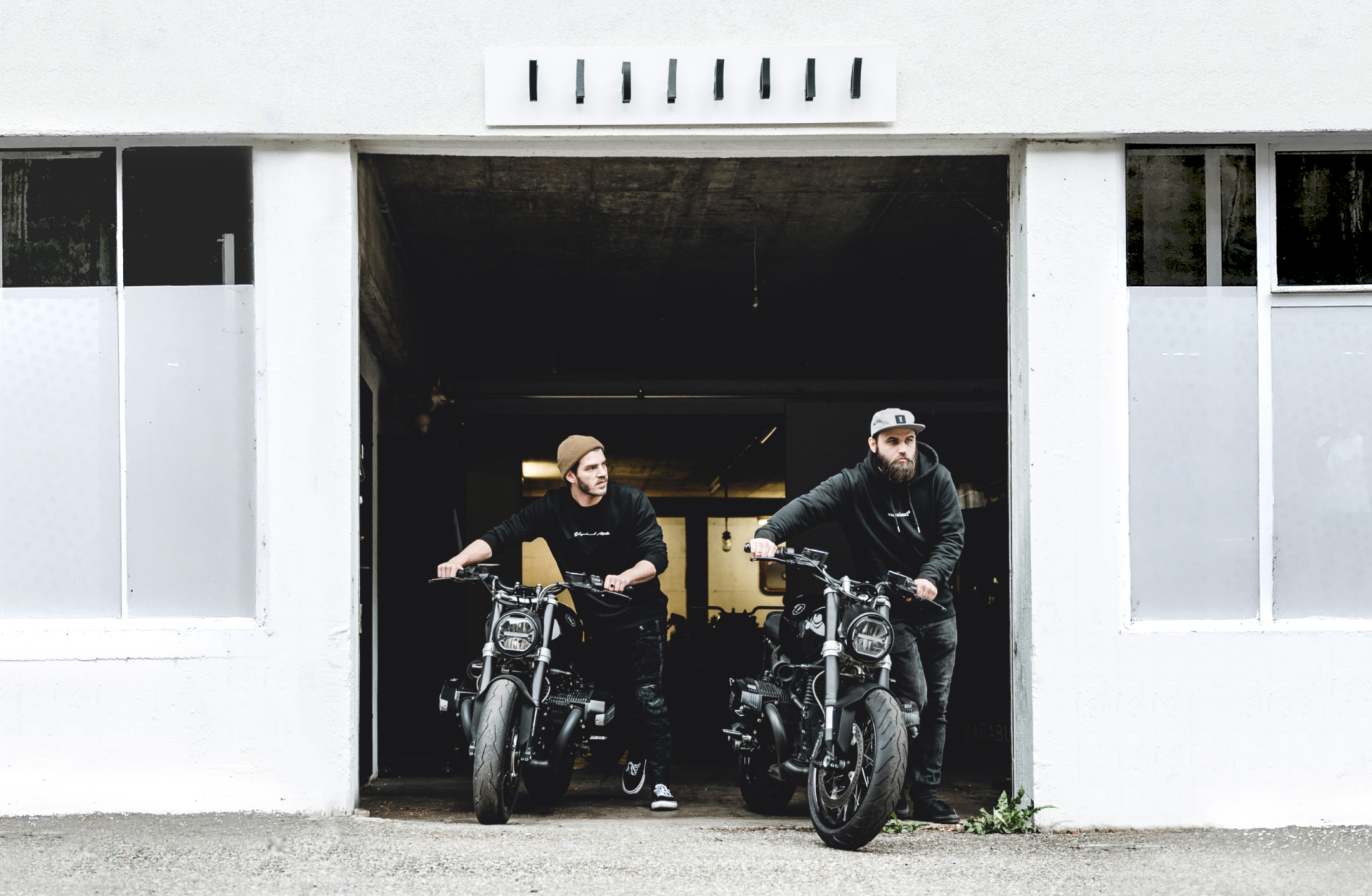 Zwei Männer sitzen auf Motorrädern vor einer weißen Garagenfassade.