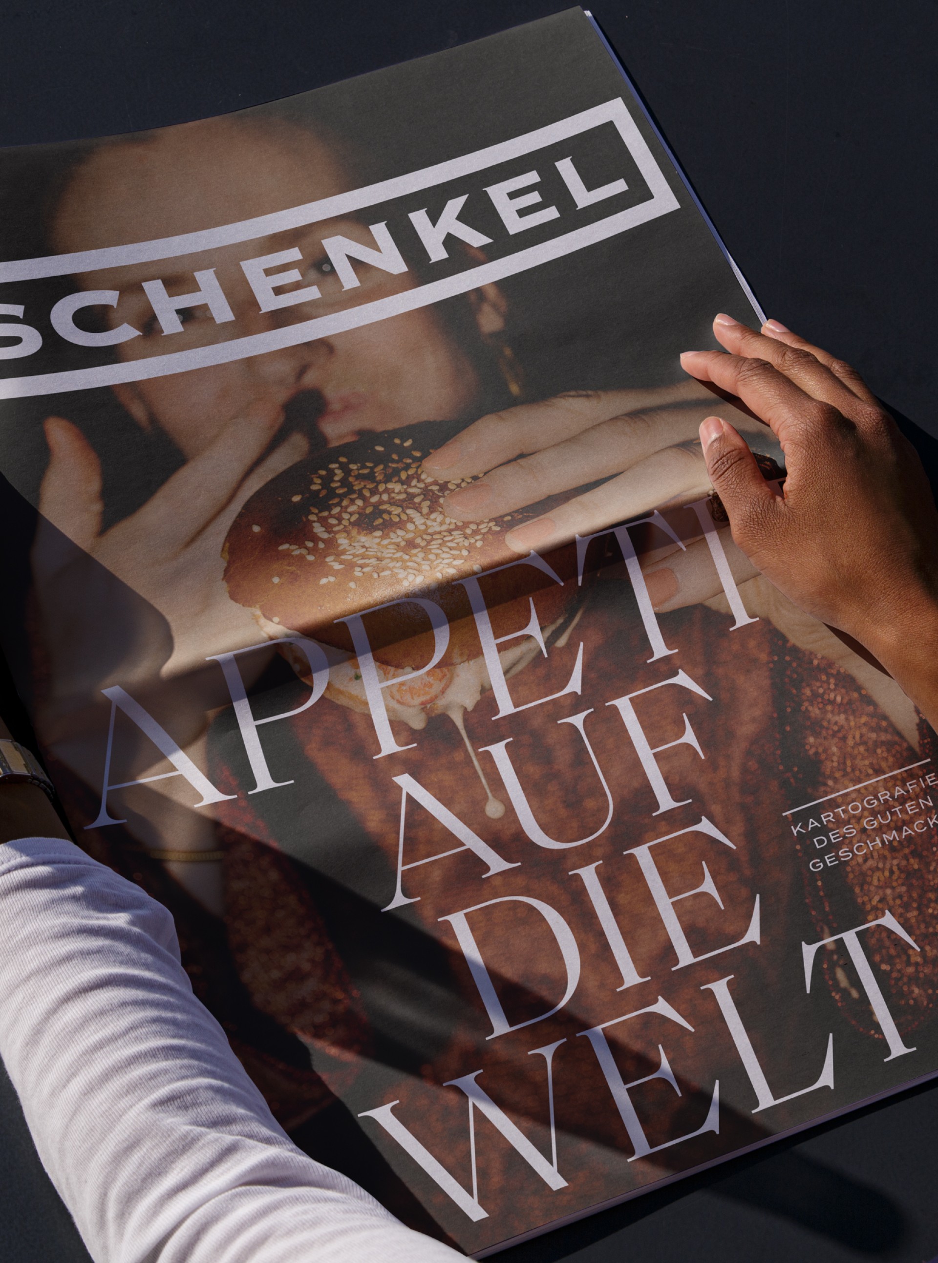 Nahaufnahme einer Zeitschrift mit dem Titel „SCHENKEL“. Das Bild zeigt eine Person, die einen Burger isst. Text: "Appetit auf die Welt".