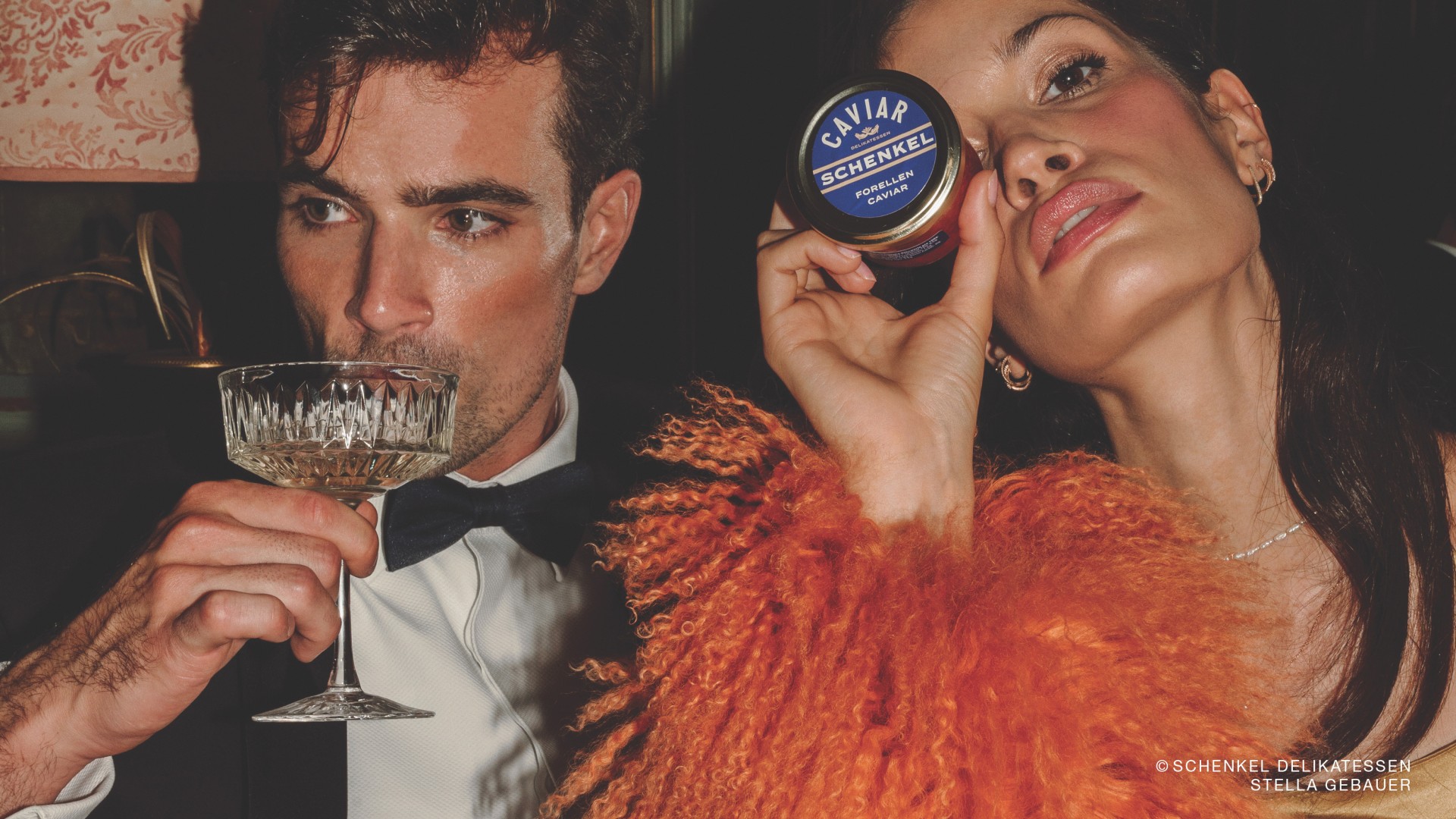 Ein Mann trinkt Champagner, eine Frau hält „Caviar Schenkel Forellen Caviar“ in der Hand.