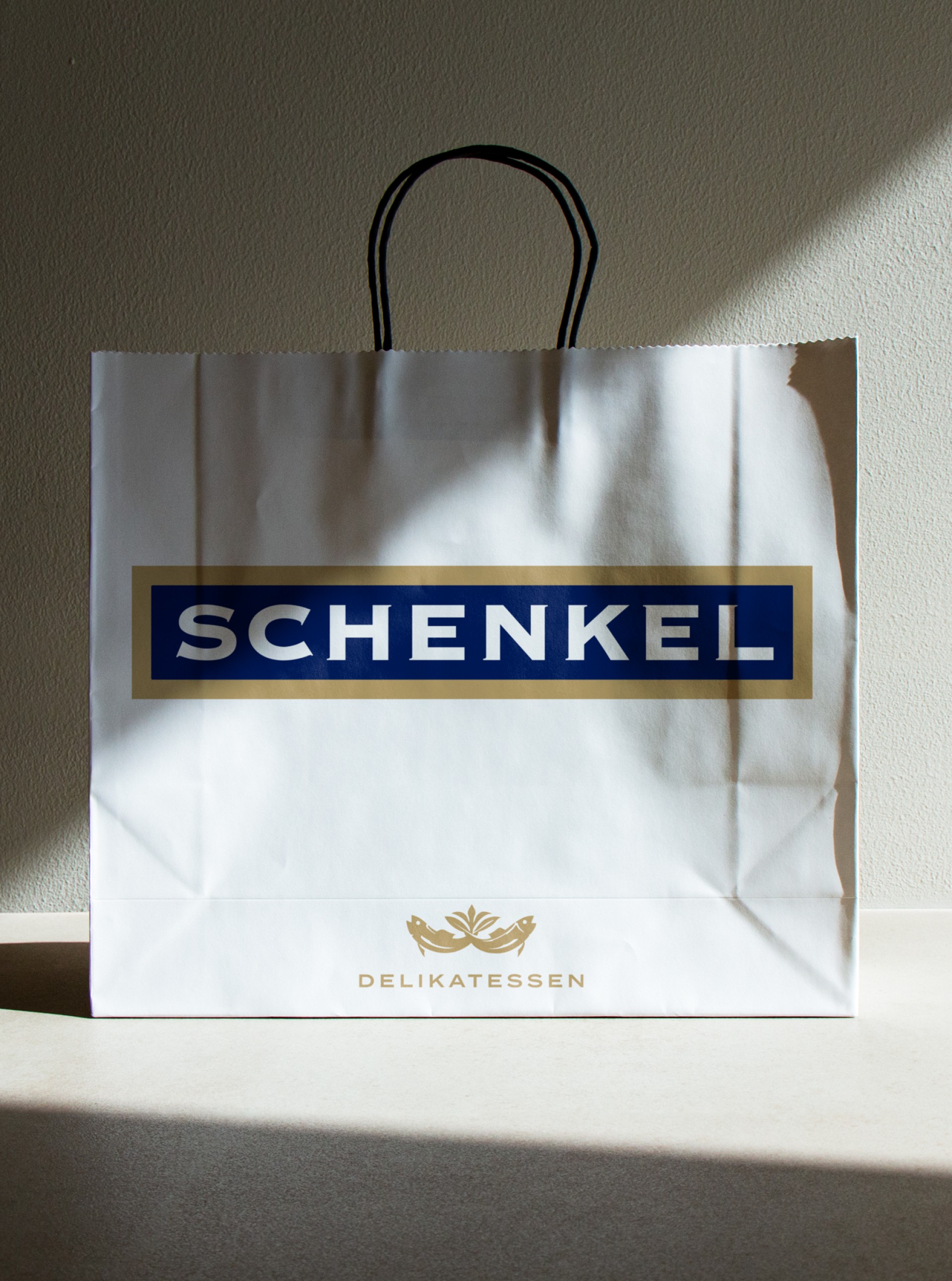 Weiße Einkaufstüte von Schenkel Delikatessen mit schwarzem Henkel und Schattenwurf.