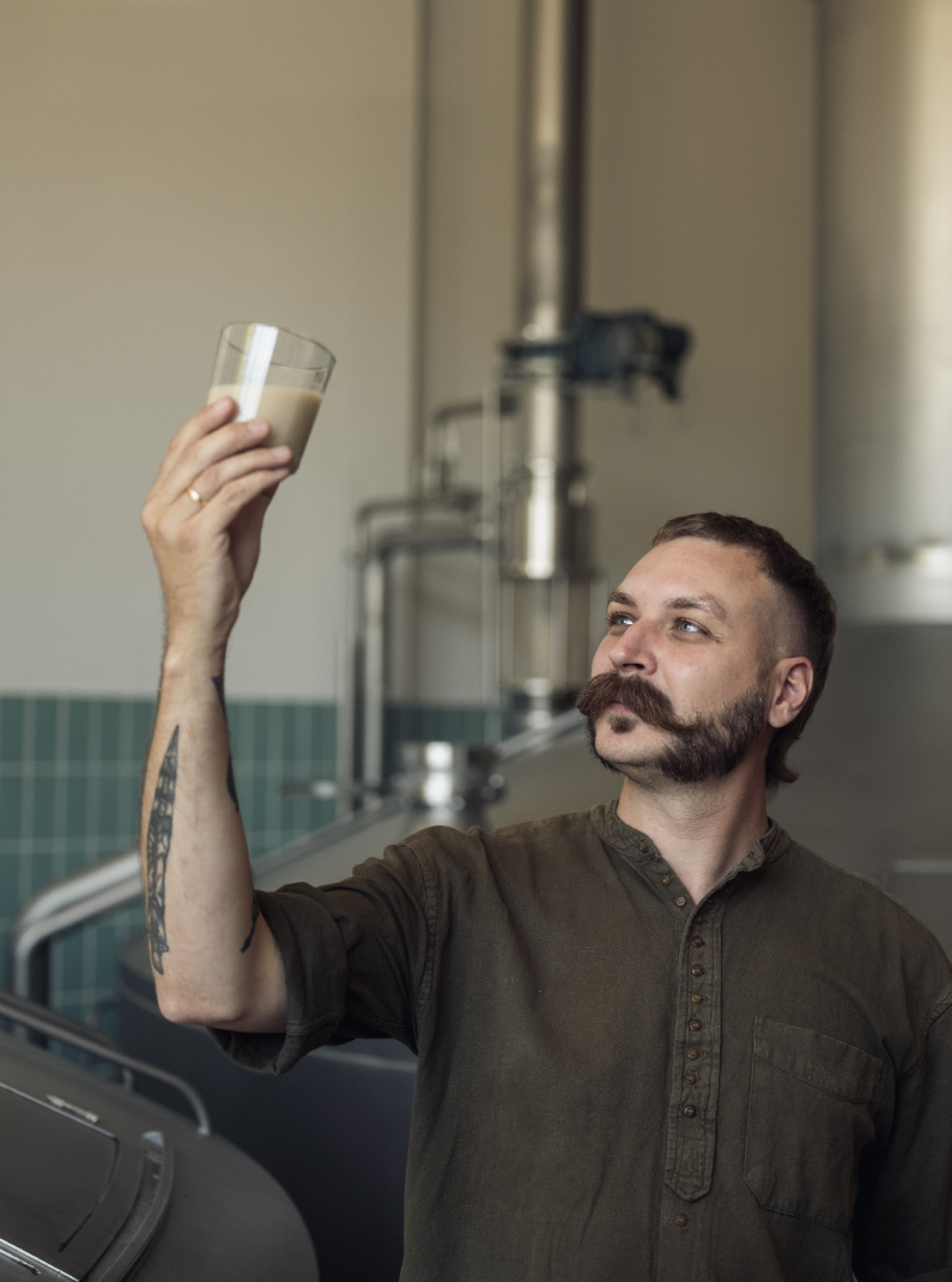 Mann mit Schnurrbart, Tätowierung, und olivgrünem Hemd, der Getränk im Glas in Brauerei betrachtet.