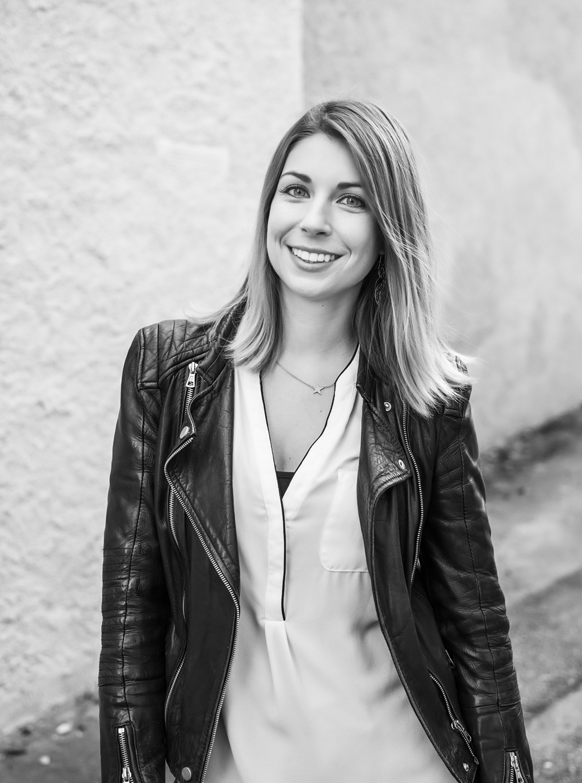 Eine lächelnde Frau mit blonden Haaren trägt eine Lederjacke und eine Bluse. Es ist ein Schwarzweißfoto.