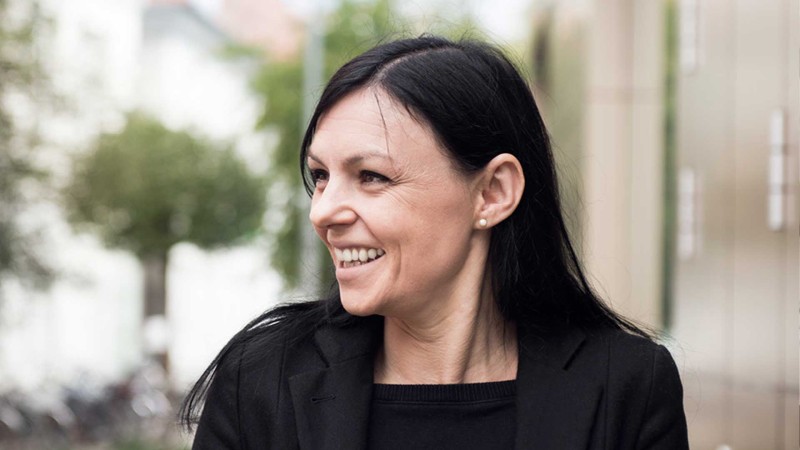 Eine Frau mit dunklen Haaren lächelt und trägt eine schwarze Jacke und ein schwarzes Hemd.