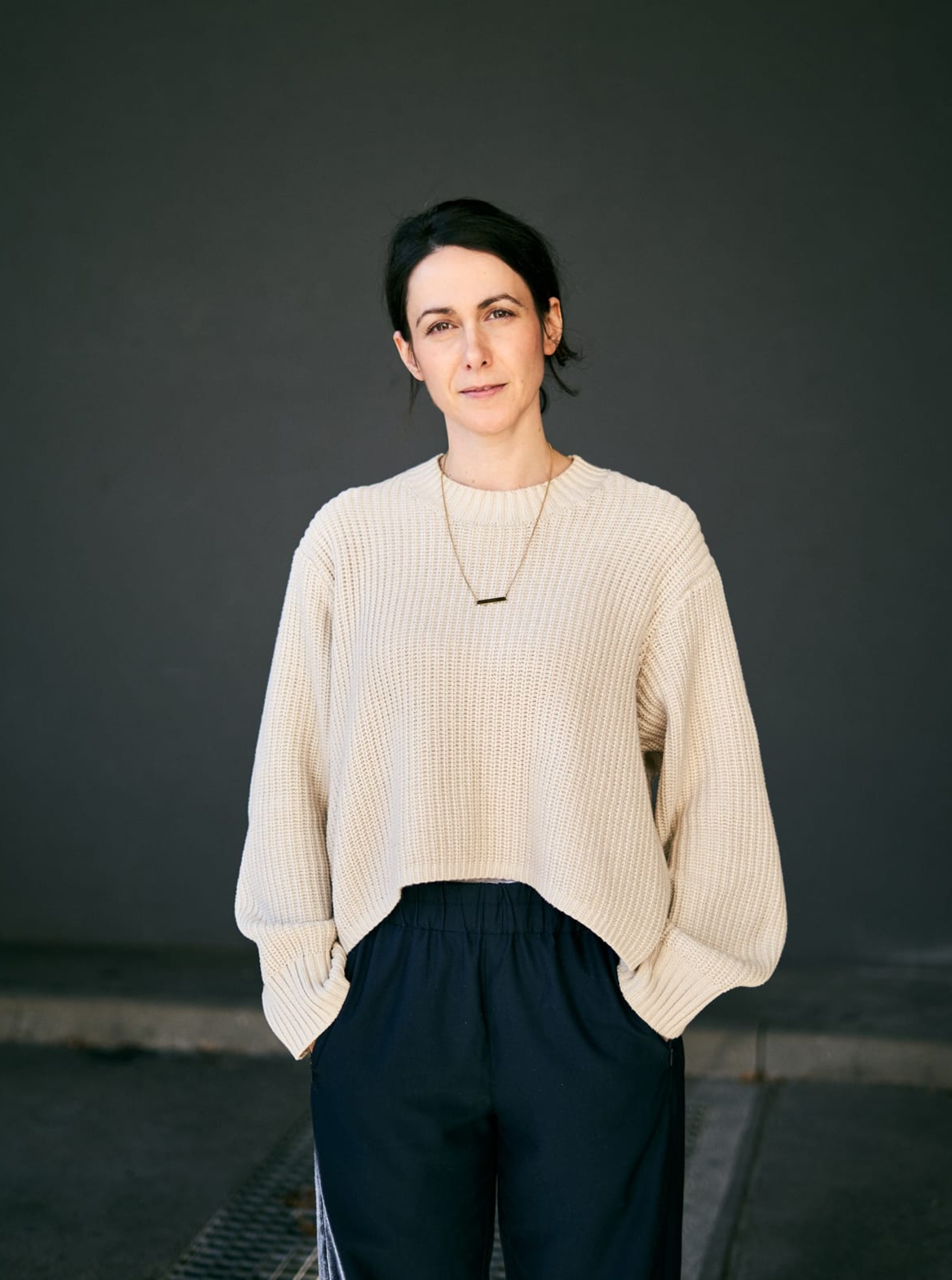 Porträt einer Frau mit dunklem Haar in einem cremefarbenen Pullover und einer dunklen Hose vor einer grauen Wand.