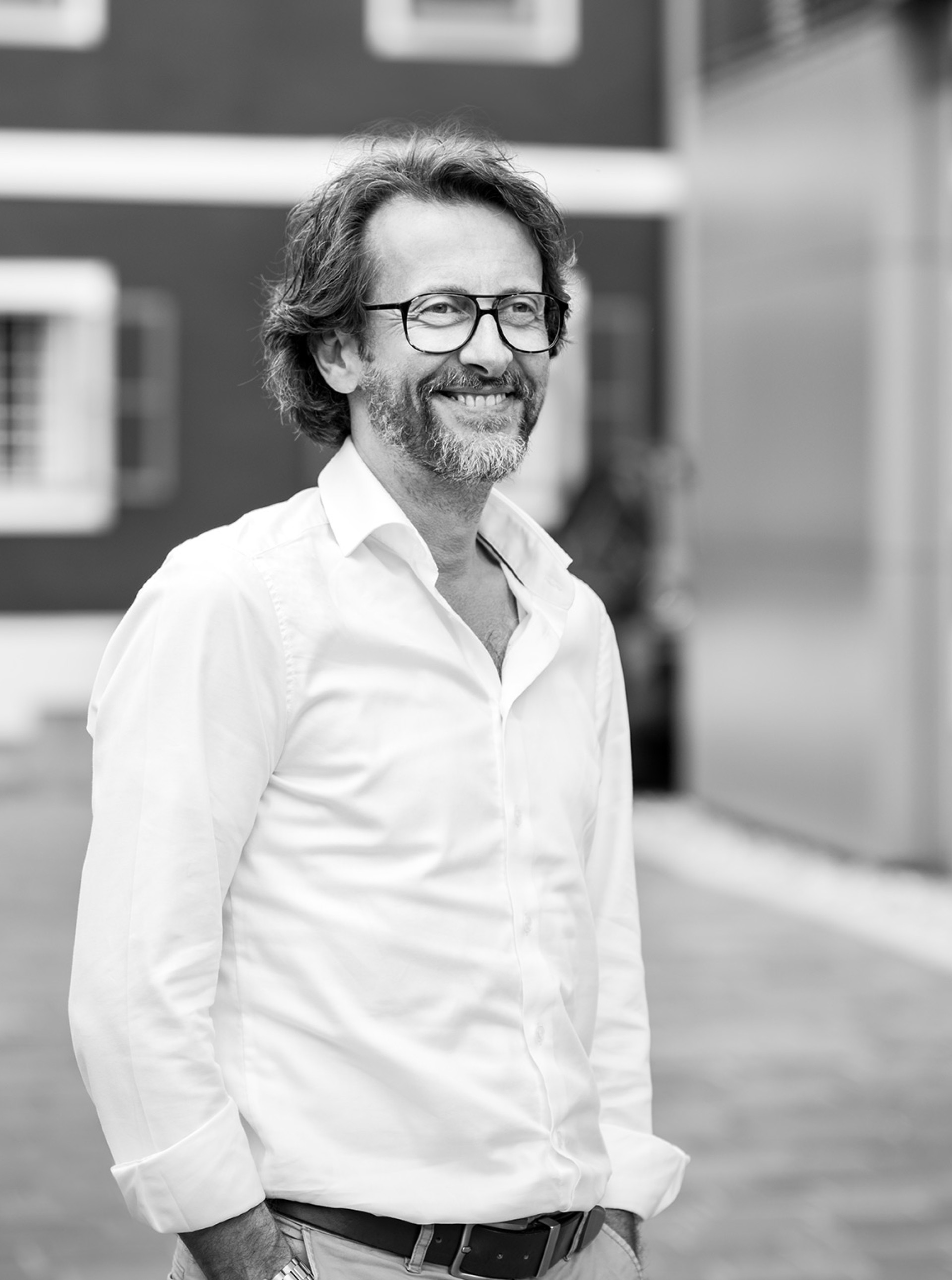 Ein Mann mit Brille und Bart, der ein weißes Hemd trägt, lächelt in Schwarzweiß.