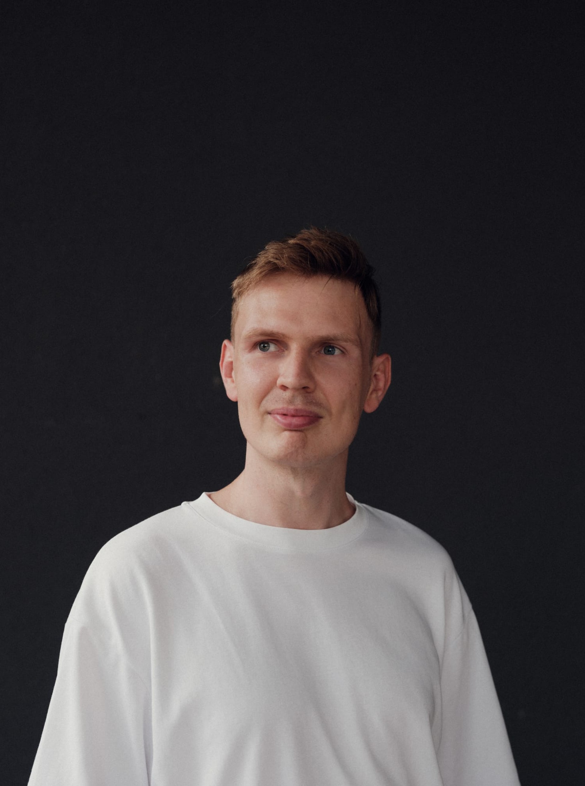Ein Mann mit kurzen roten Haaren und weißem T-Shirt vor einem schwarzen Hintergrund.