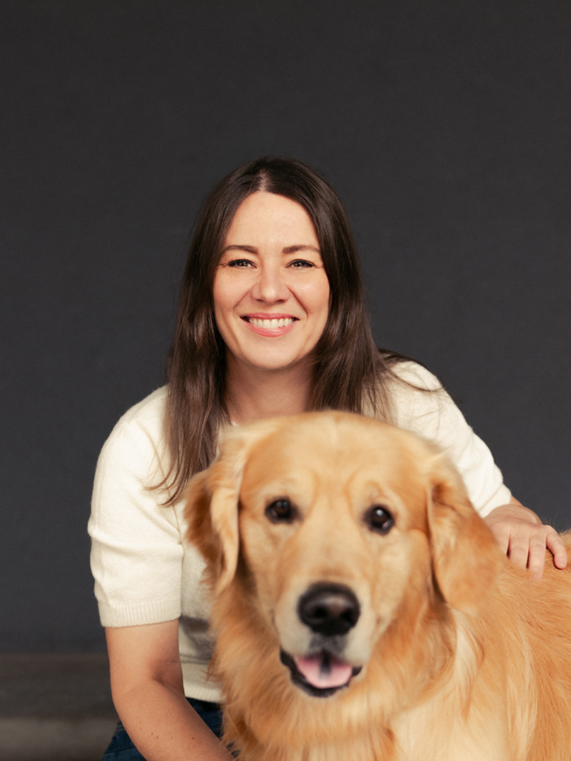 Eine lächelnde Frau mit einem Golden Retriever vor einem grauen Hintergrund.