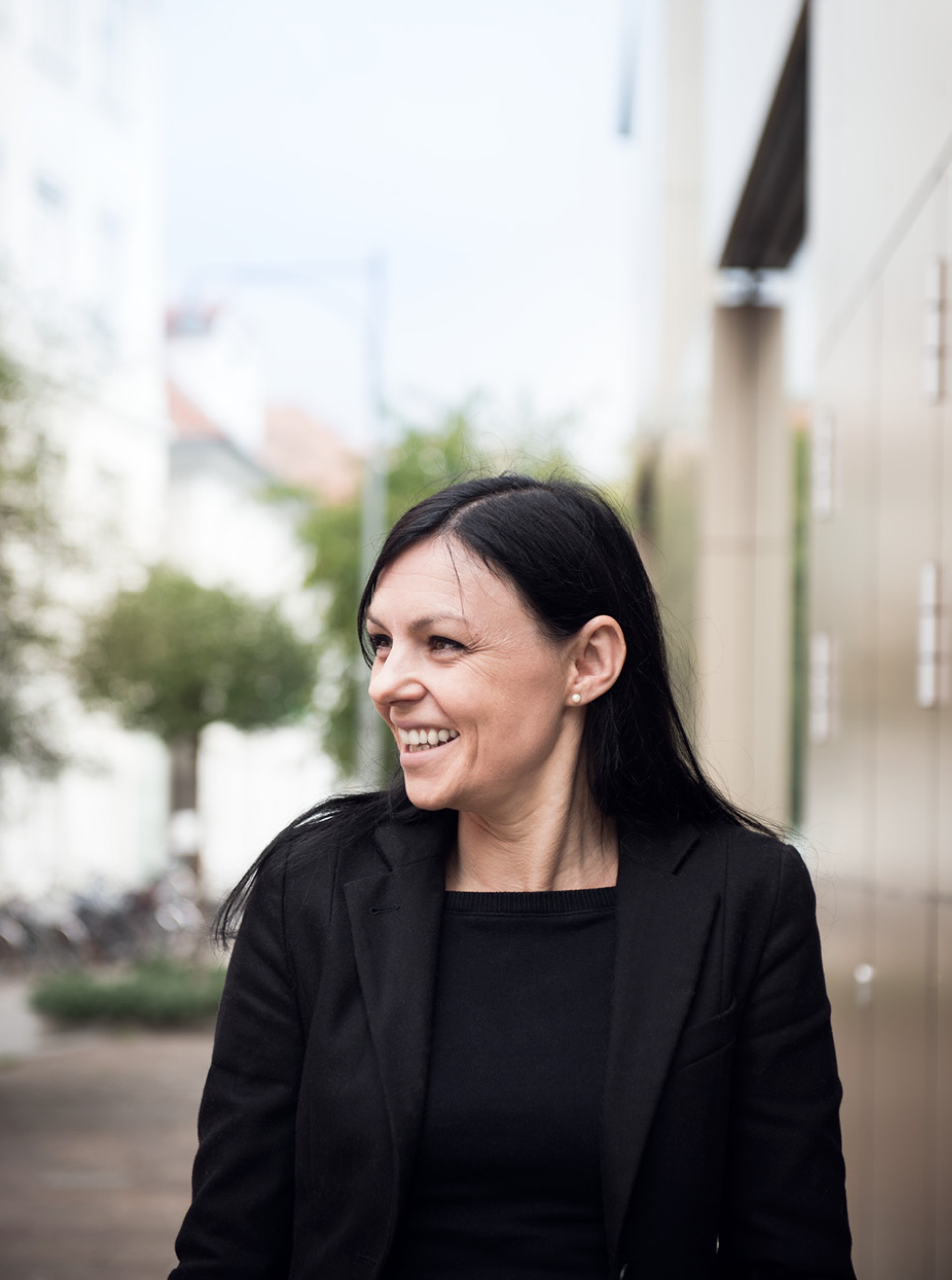 Eine lachende Frau mit dunklem Haar, in einem schwarzen Blazer, blickt zur Seite, im Freien.