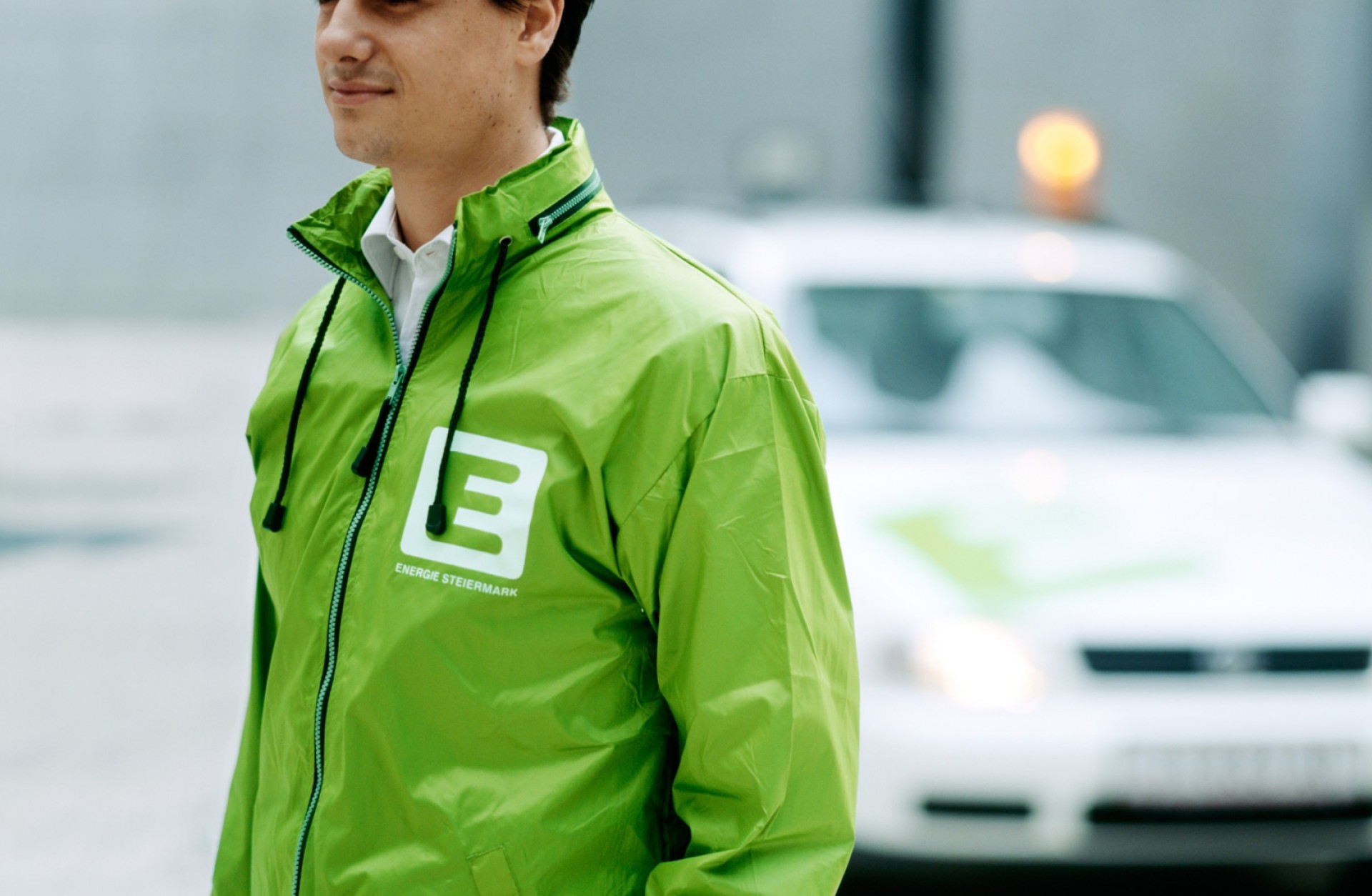 Ein Mann trägt eine grüne Jacke mit dem Logo von Energie Steiermark. Im Hintergrund ist ein Auto.