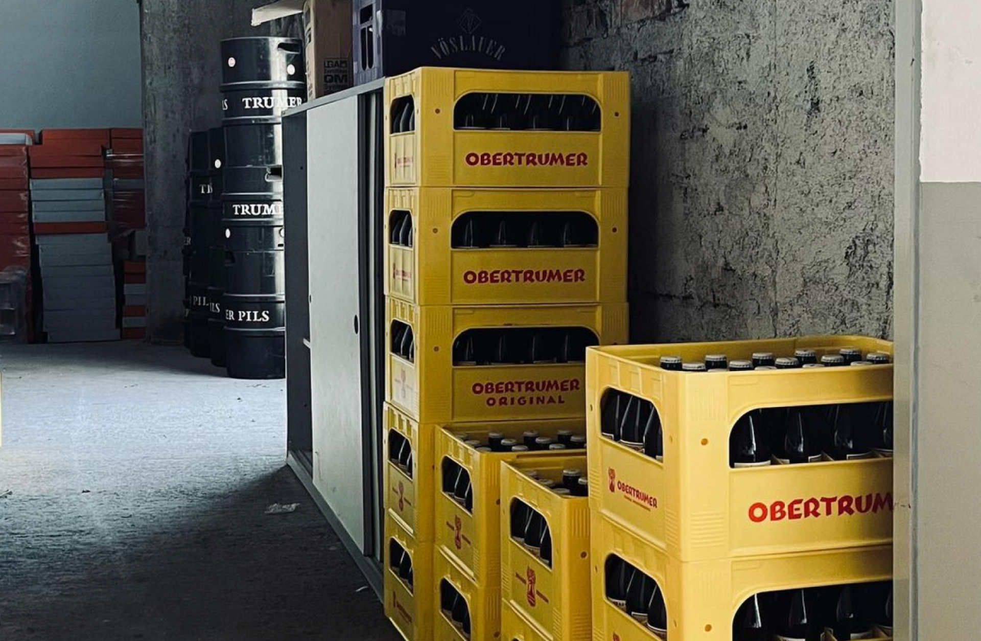 Stapel von Bierfässern "Trumer Pils" und gelben Kisten "Obertumer Original" in einem Lagerraum.
