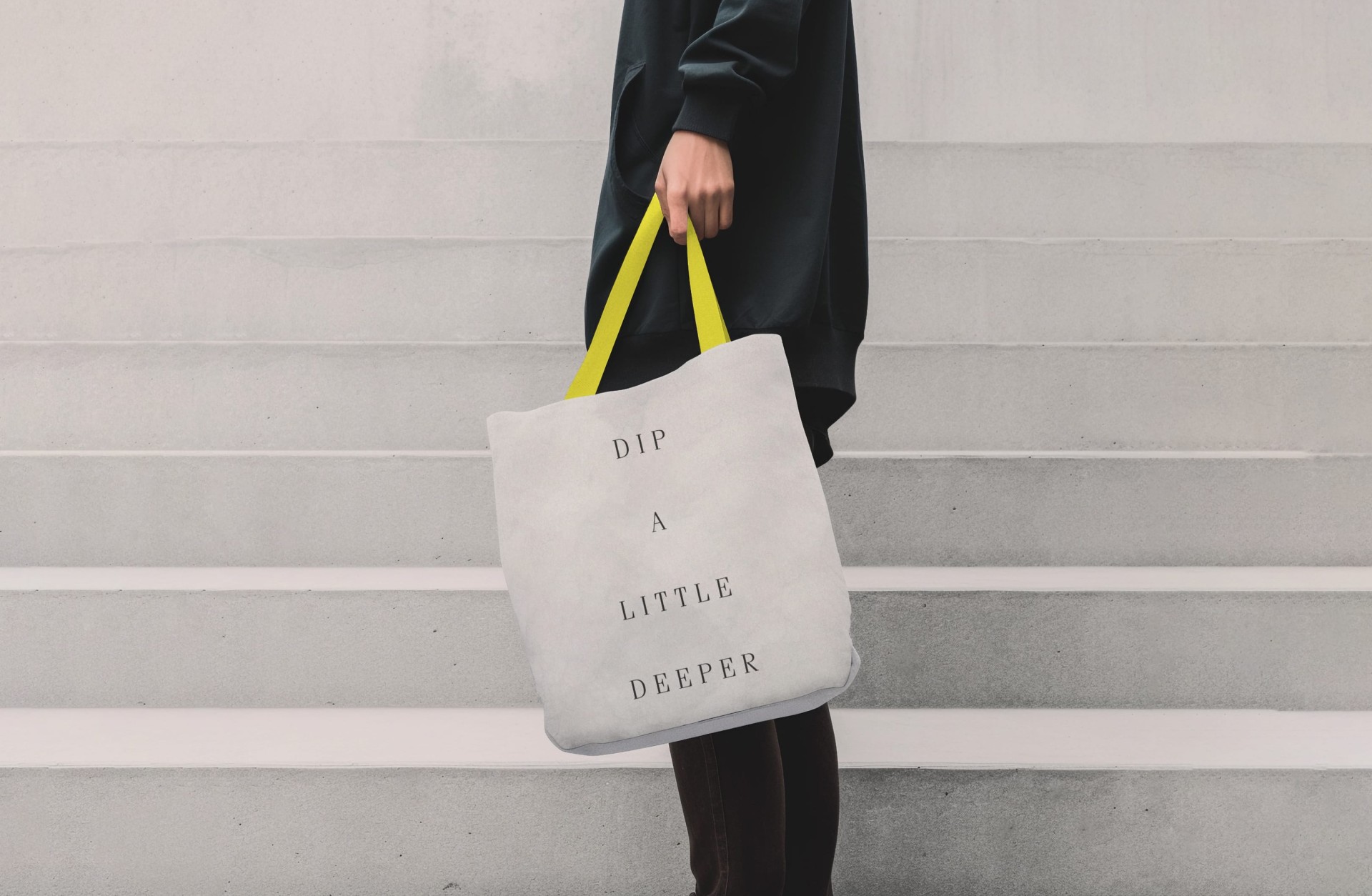 Person hält Tasche mit gelben Trägern und dem Text "DIP A LITTLE DEEPER" vor einer Treppe.